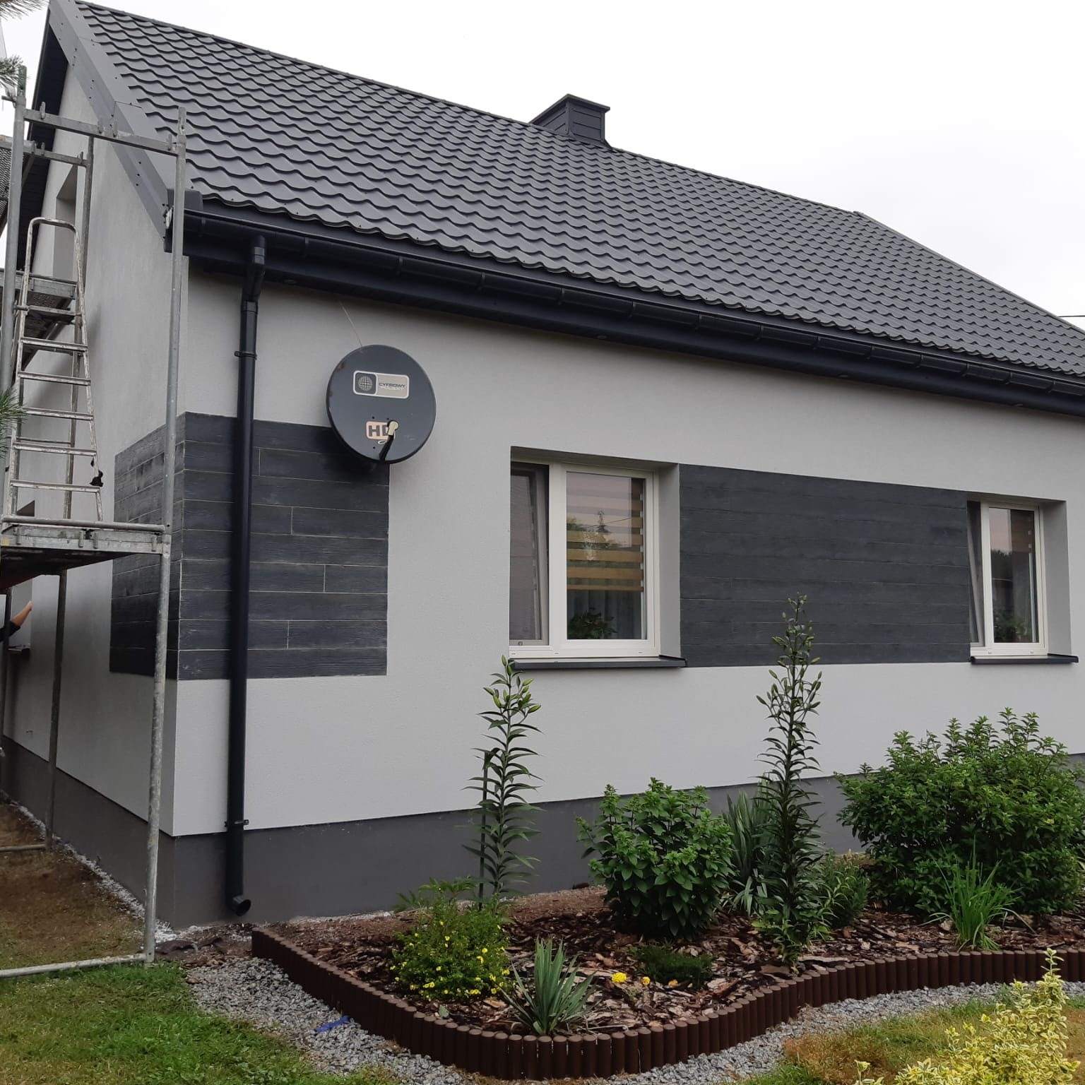 Dom z szarą elewacją i ciemnymi panelami imitującymi drewno, widoczne rusztowanie, ogródek z roślinami i kwiatami. Nowoczesny design.