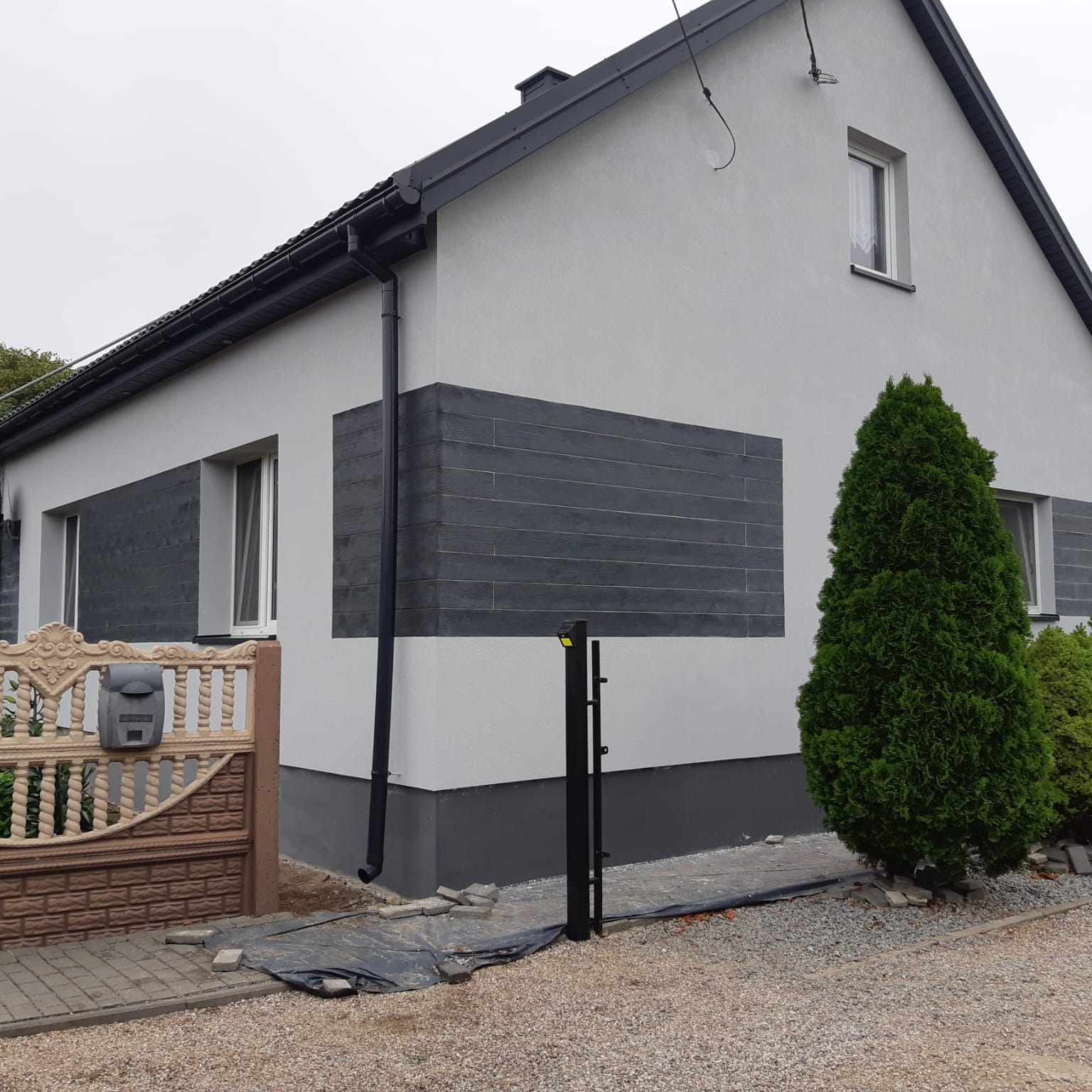 Wykończona elewacja domu jednorodzinnego z szarymi panelami imitującymi drewno, widok z zewnątrz na tle szarego nieba, z ozdobnym ogrodzeniem i tujami.