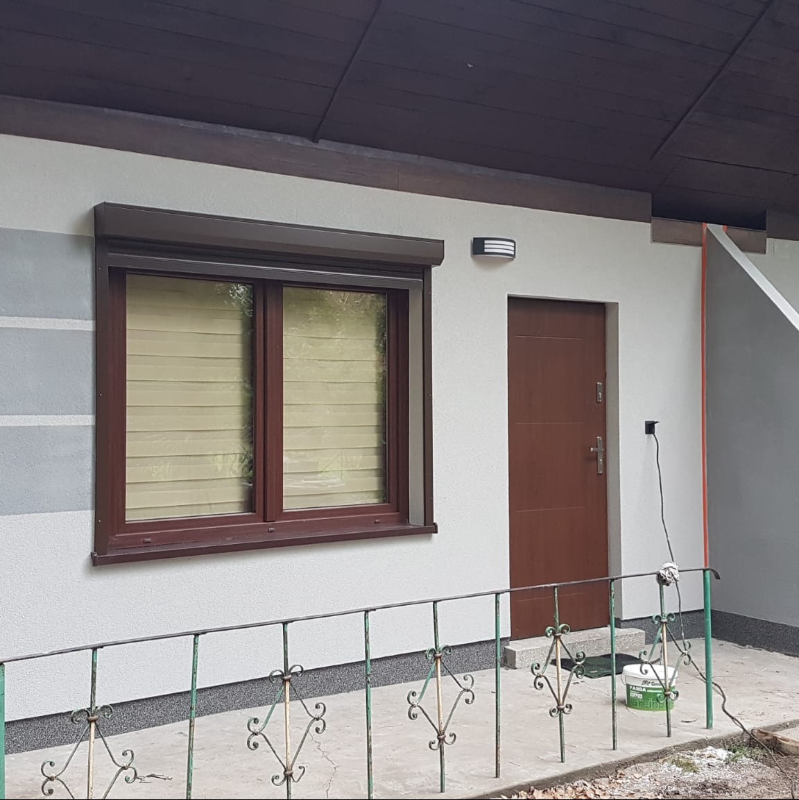 Elewacja domu po renowacji: okno z roletą, brązowe drzwi wejściowe, świeżo pomalowana ściana w odcieniach szarości, dekoracyjny płotek.