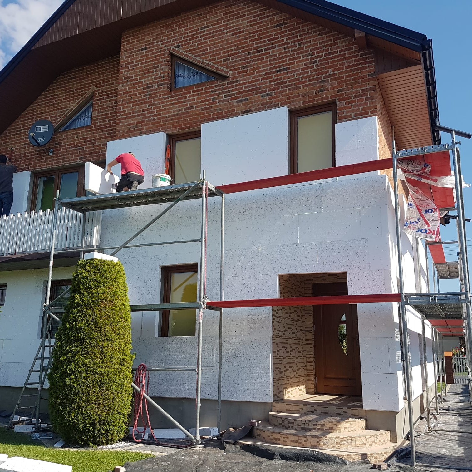 Ocieplanie elewacji domu jednorodzinnego z cegły. Prace na rusztowaniu, widoczne płyty styropianowe i pracownicy. Częściowo ocieplona ściana frontowa.