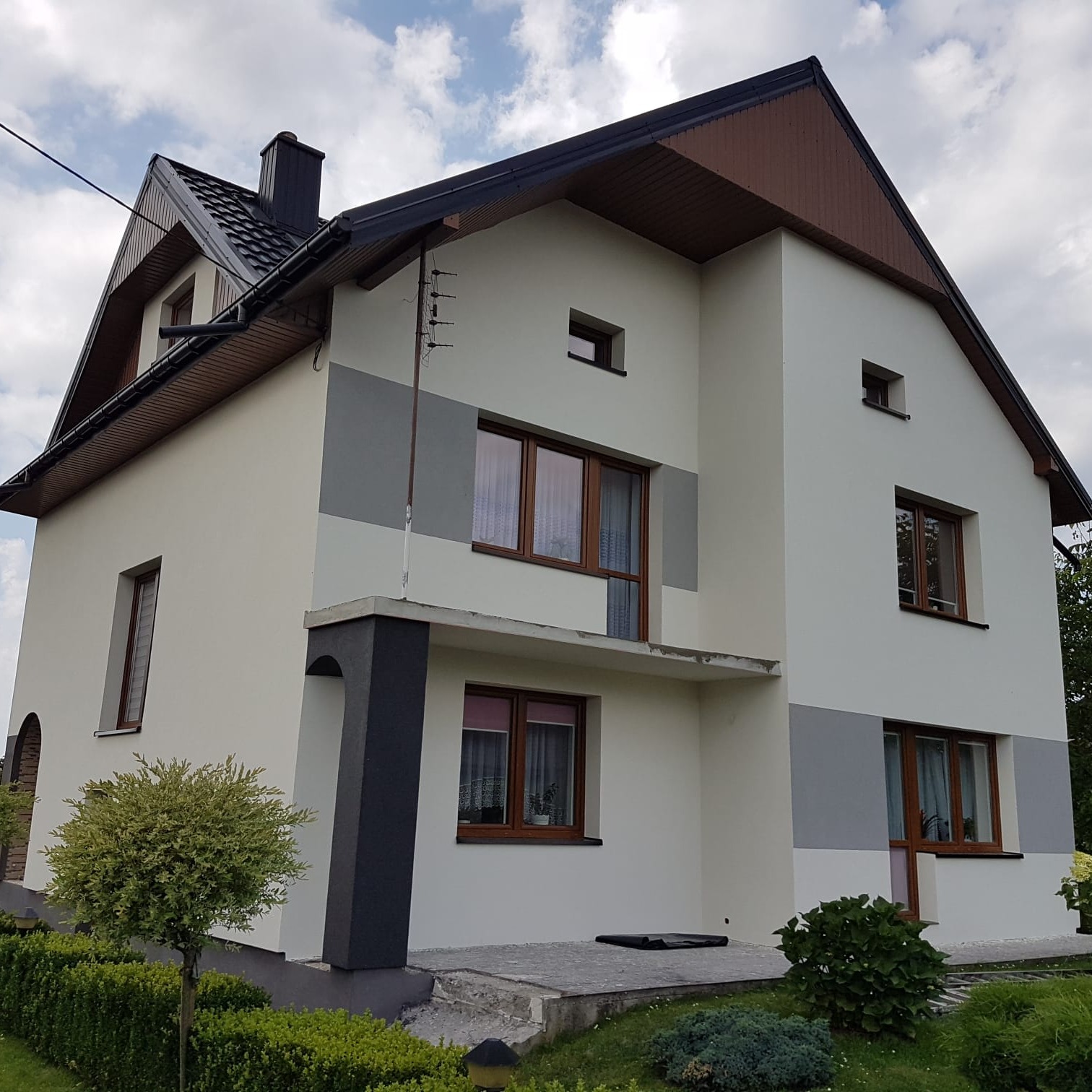 Dwukondygnacyjny dom z jasną elewacją, ciemnymi akcentami i brązowymi oknami. Widoczne fragmenty szarego pasa na elewacji. Uwagę przykuwa antena na dachu i zadbana zieleń.