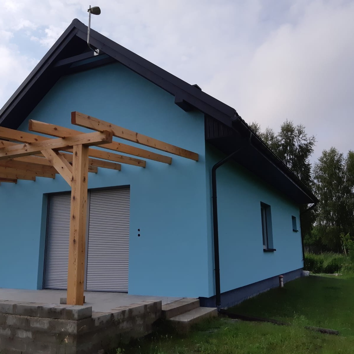 Błękitny dom jednorodzinny z drewnianą pergolą, czarnym dachem i szarymi roletami zewnętrznymi. Widok z boku, trawnik przed budynkiem.