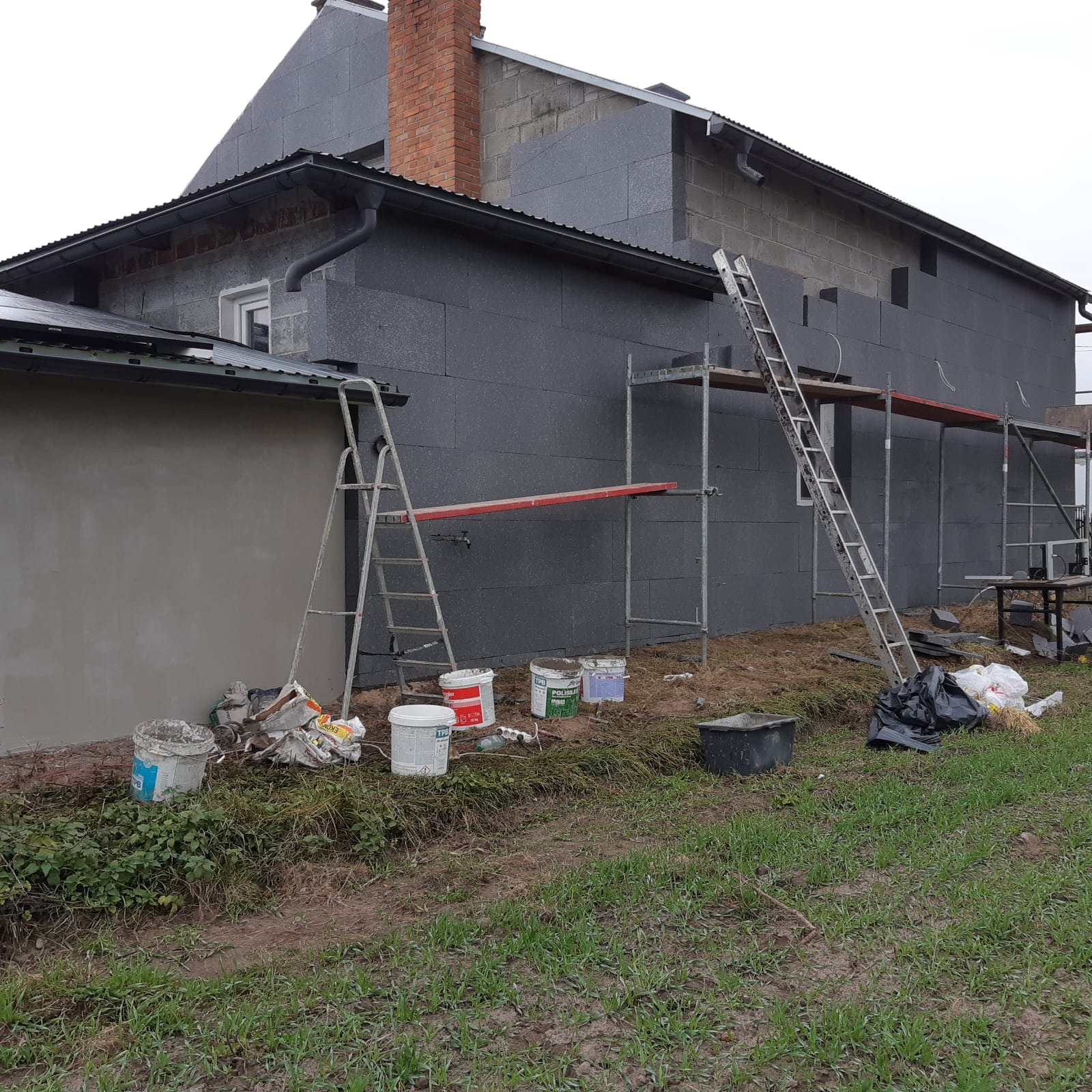 Dom w trakcie ocieplania elewacji styropianem grafitowym. Widoczne rusztowania, drabiny i pojemniki z zaprawą. Częściowo ocieplony budynek z ceglanym kominem.