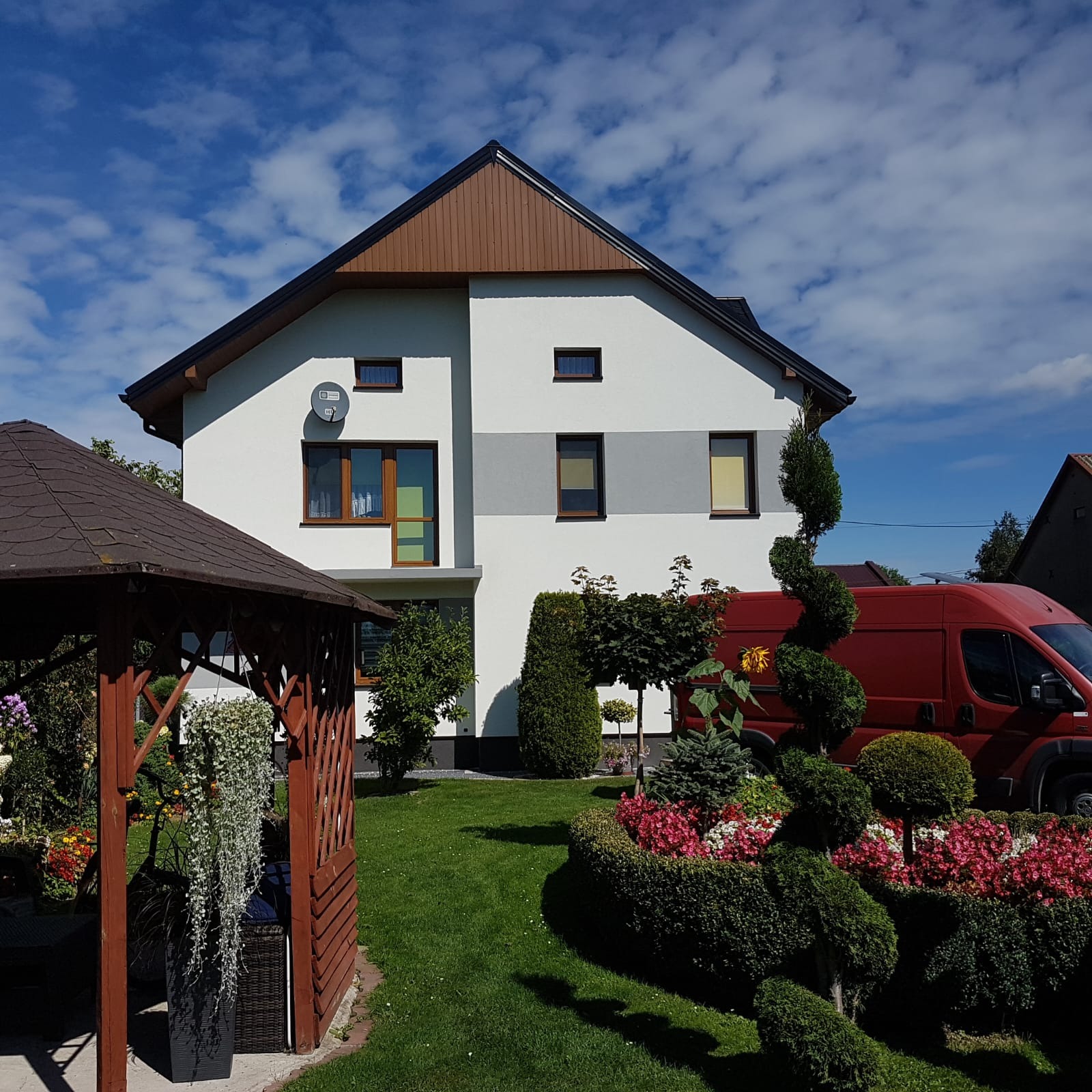 Dwukondygnacyjny dom z biało-szarą elewacją, brązowym dachem i oknami, otoczony zadbanym ogrodem z altaną i czerwonym busem w tle, widok z poziomu gruntu.