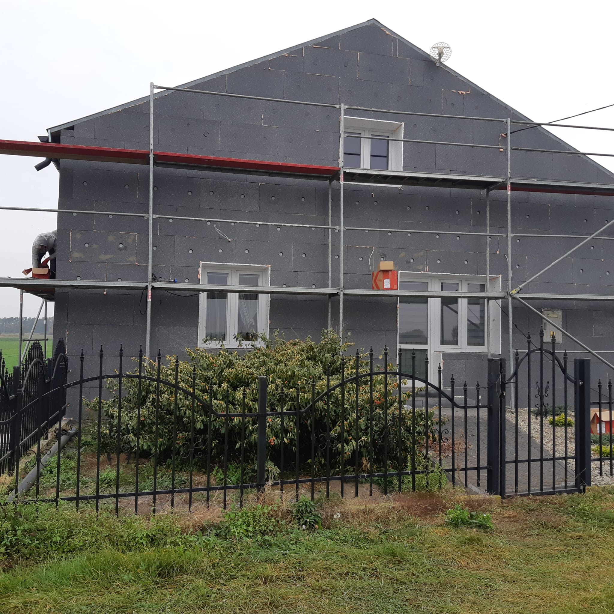 Dom w trakcie ocieplania styropianem grafitowym, widoczne rusztowania i pracownik na nich. Ogród z czarnym ogrodzeniem na pierwszym planie. Etap prac.