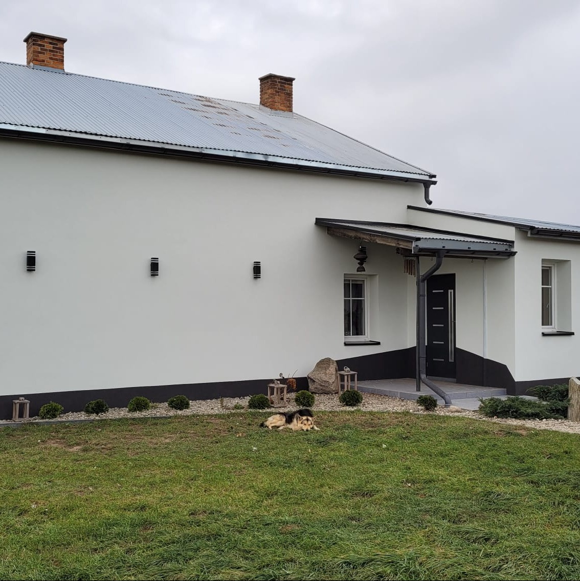 Elewacja domu jednorodzinnego w odcieniach bieli i szarości, z widocznym dachem z blachy i elementami dekoracyjnymi. Pies leży na trawniku przed budynkiem.