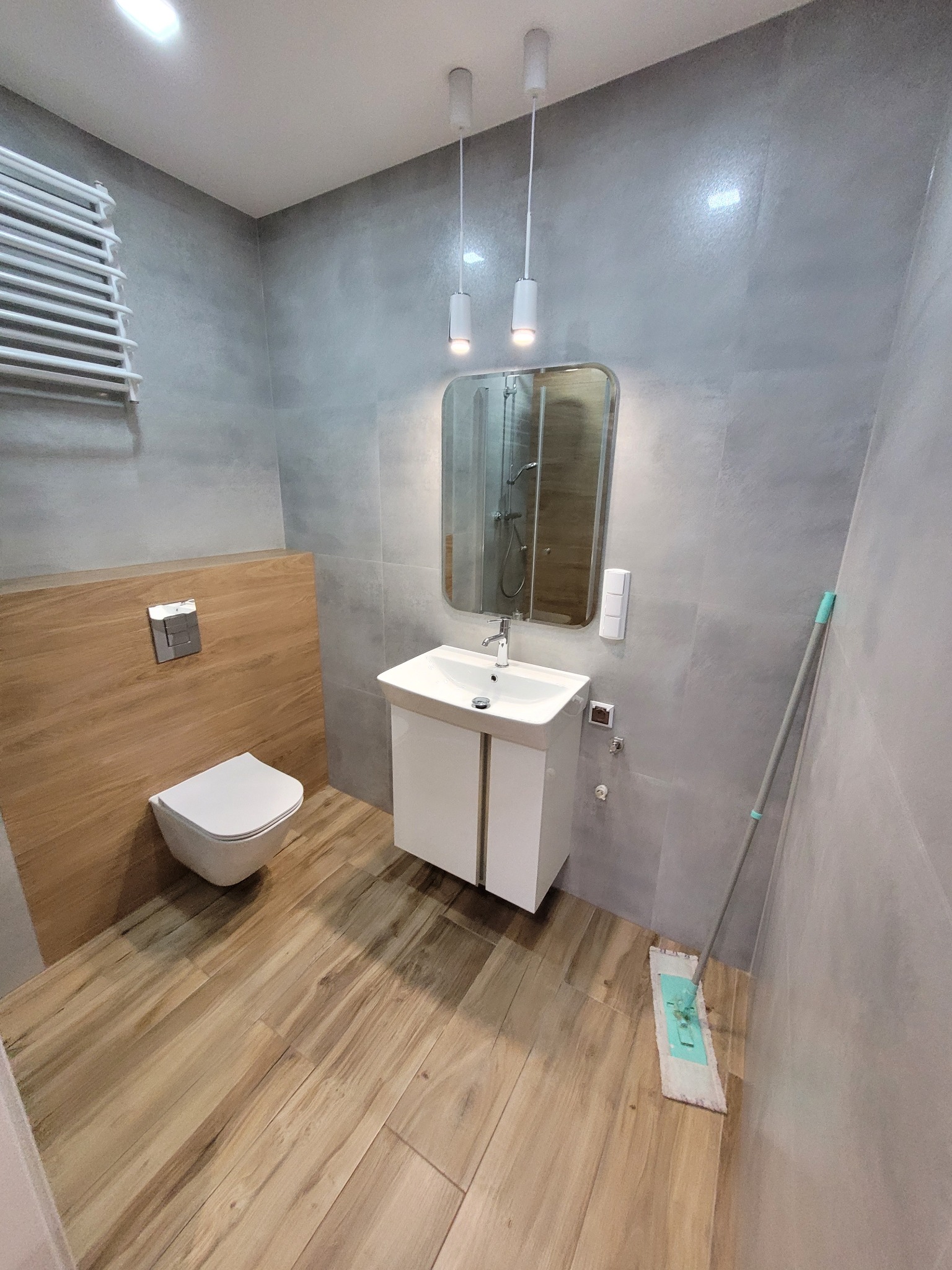 Wykończona łazienka z drewnopodobną podłogą i szarą glazurą. Umywalka z szafką, toaleta podwieszana i grzejnik drabinkowy. Minimalistyczne oświetlenie i mop przy ścianie.