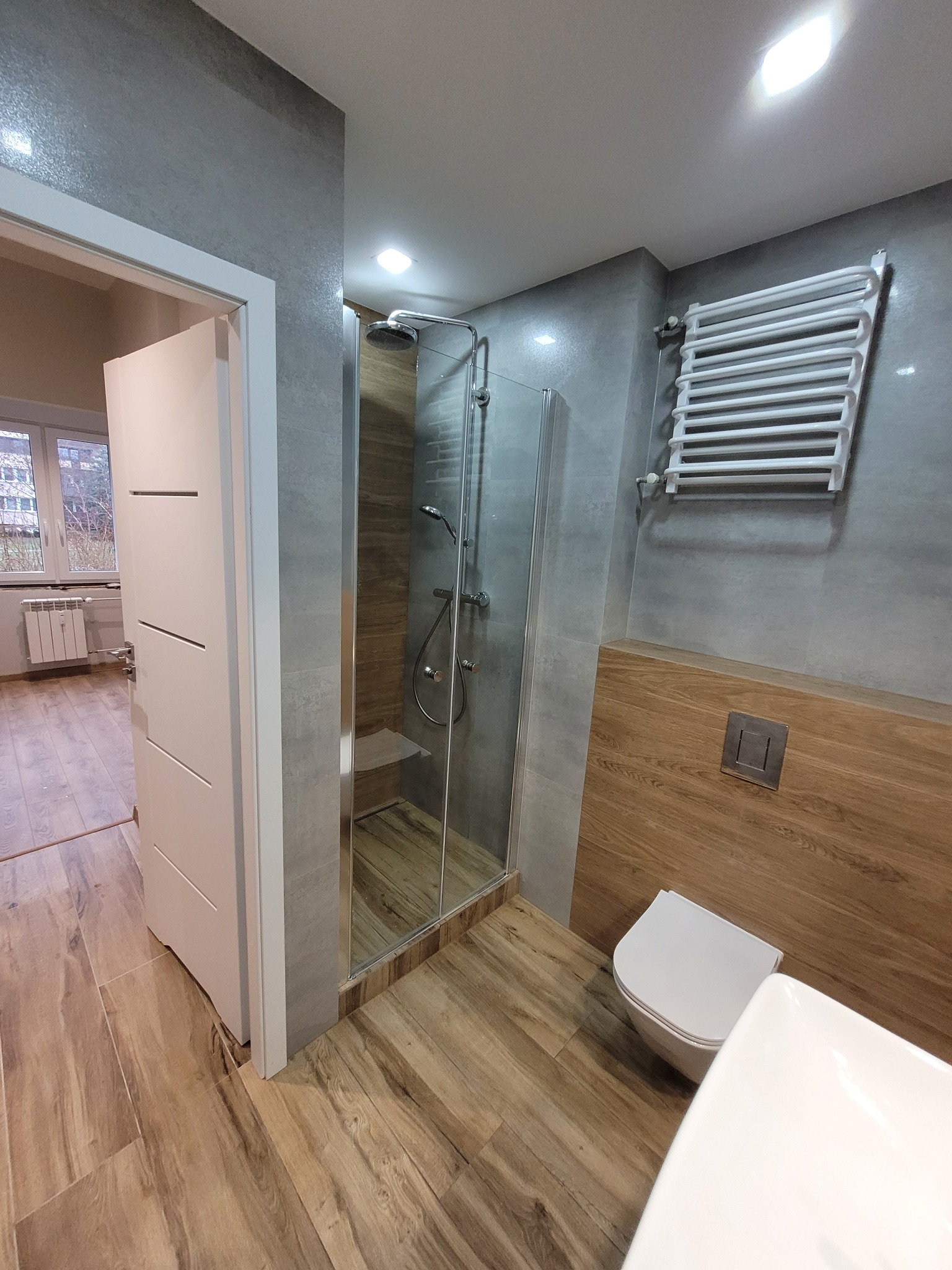 Nowoczesna łazienka po remoncie: kabina prysznicowa ze szklanymi drzwiami, drewnopodobne płytki, grzejnik drabinkowy i biała armatura. Widok zza otwartych drzwi.