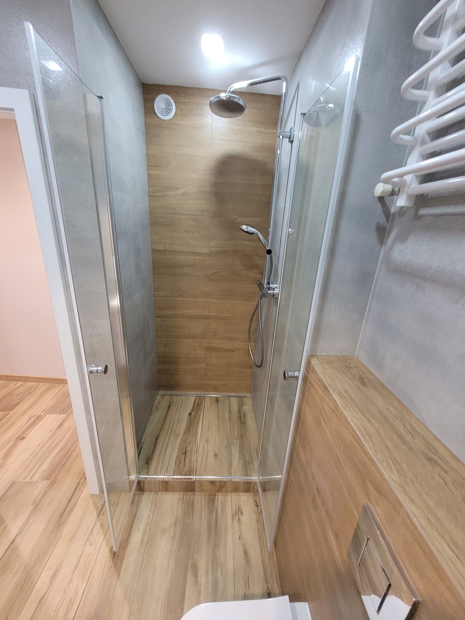 Nowoczesna kabina prysznicowa z drewnopodobnymi płytkami na ścianach i podłodze. Szklane drzwi kabiny z chromowanymi uchwytami. Grzejnik drabinkowy na ścianie.