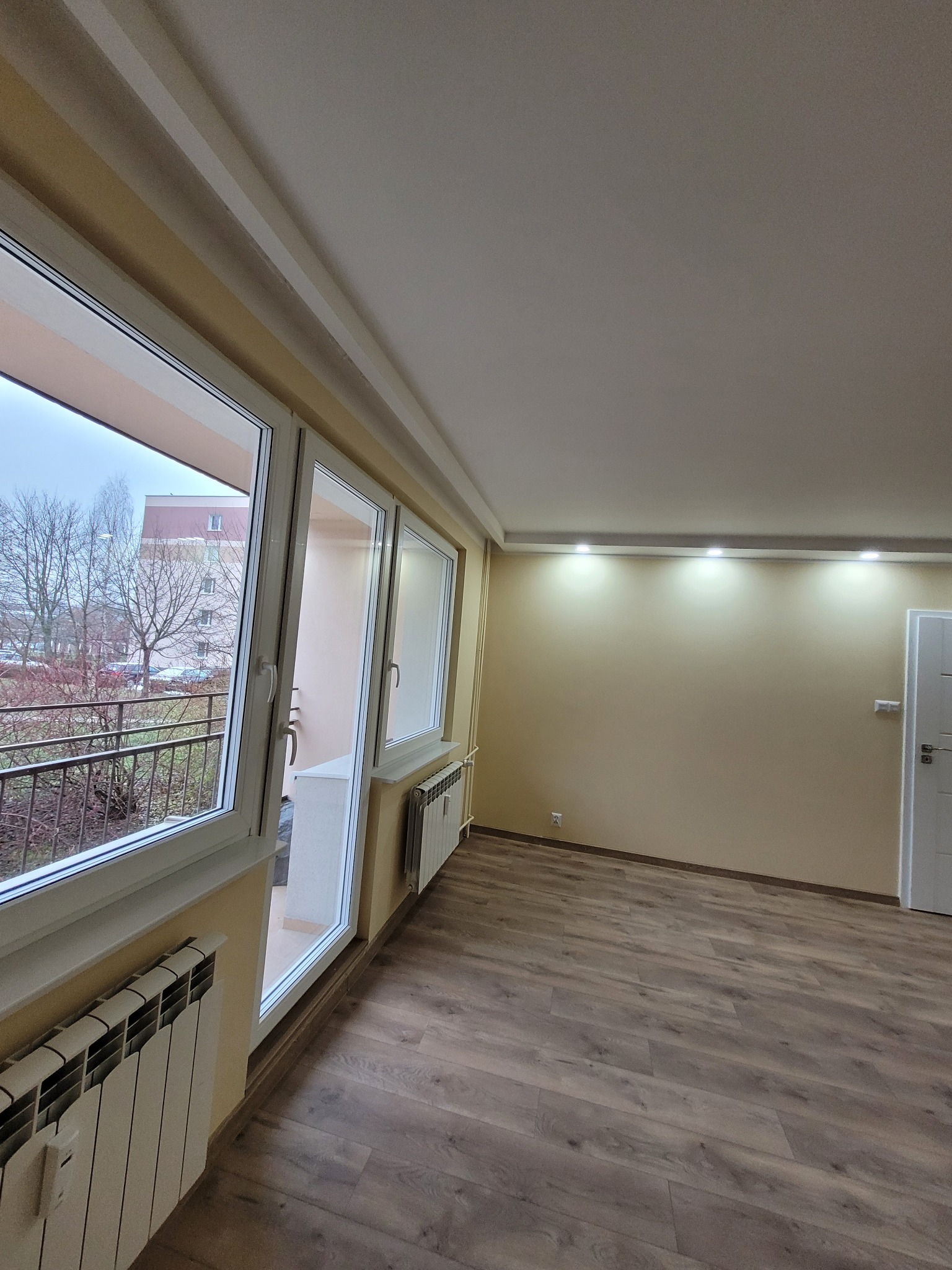 Jasne wnętrze po remoncie: parkiet, ściany w odcieniach beżu, białe okna z widokiem na balkon i drzewa. Grzejniki pod oknami, sufit z oświetleniem punktowym.
