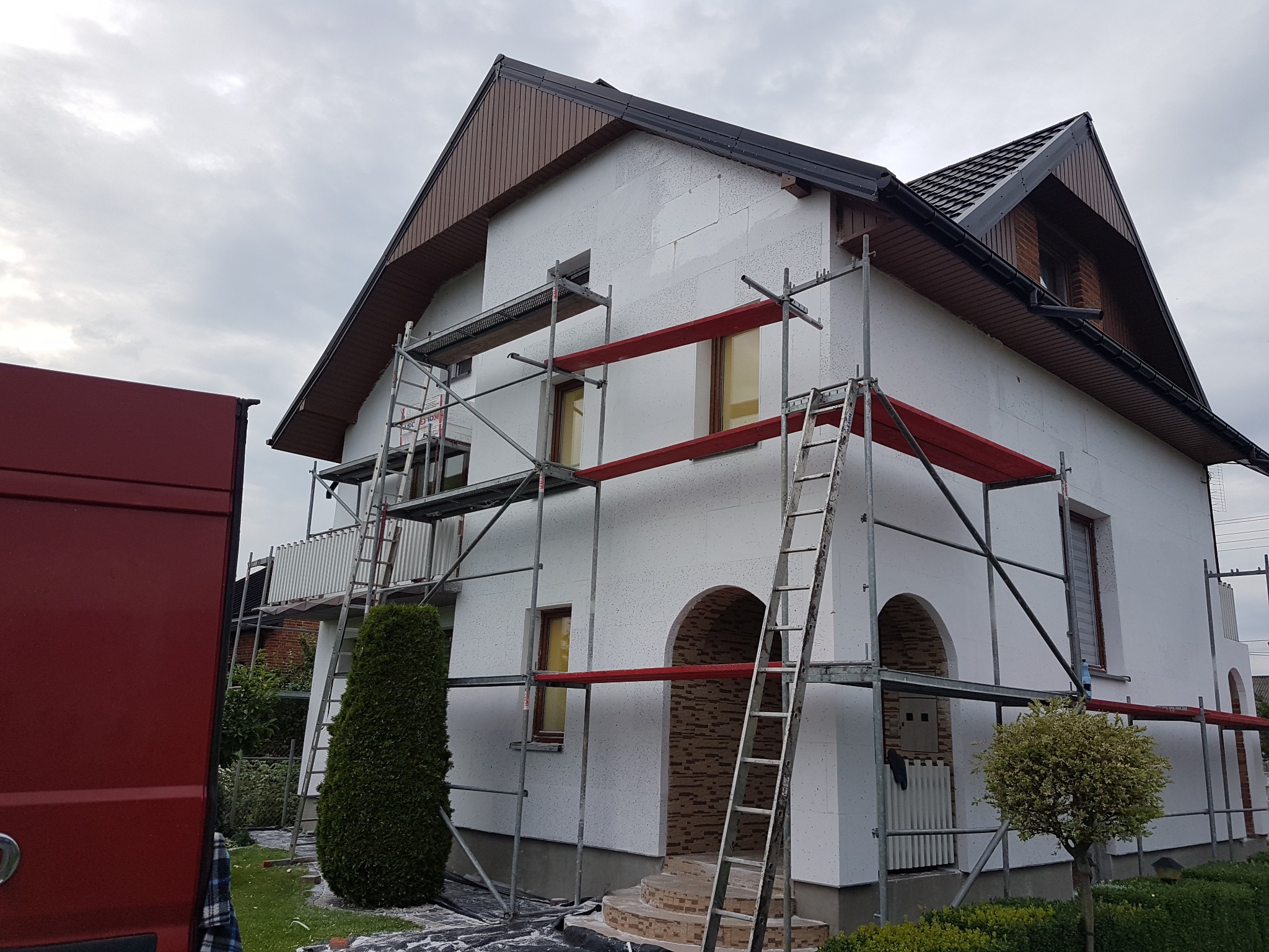 Dom w trakcie ocieplania styropianem, widoczne rusztowania i drabina. Częściowo ocieplona elewacja z widocznymi elementami dekoracyjnymi. Czerwony samochód dostawczy z lewej.