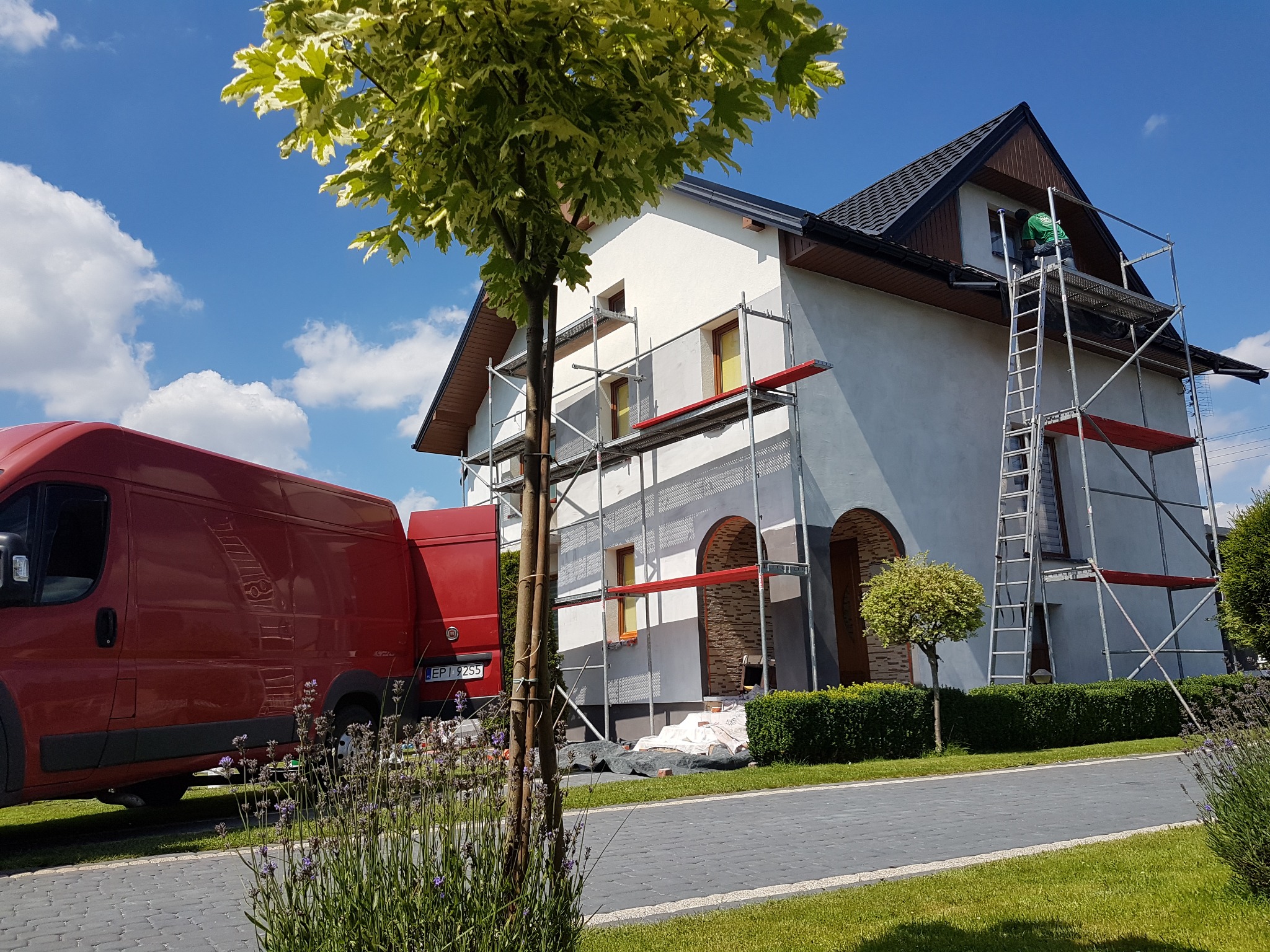 Dom w trakcie prac elewacyjnych: rusztowania, pracownik przy oknie, częściowo otynkowana ściana, czerwony bus w tle, błękitne niebo z chmurami.