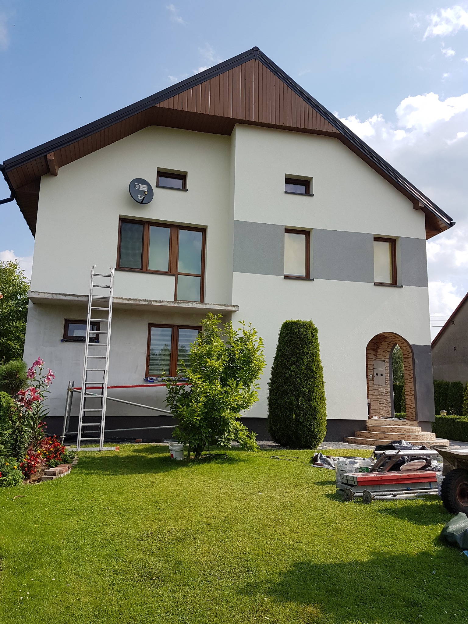 Dwukondygnacyjny dom po częściowym malowaniu elewacji w Grabicy. Widoczne rusztowanie, drabina i narzędzia malarskie na trawniku. Świeża, jasna kolorystyka.