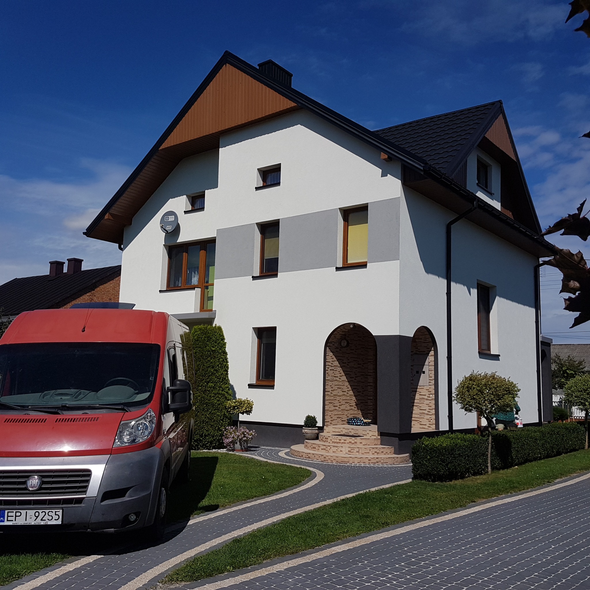 Dwukondygnacyjny dom z białą elewacją, szarymi akcentami i brązowym dachem, widziany z poziomu chodnika. Czerwony samochód dostawczy zaparkowany z przodu.