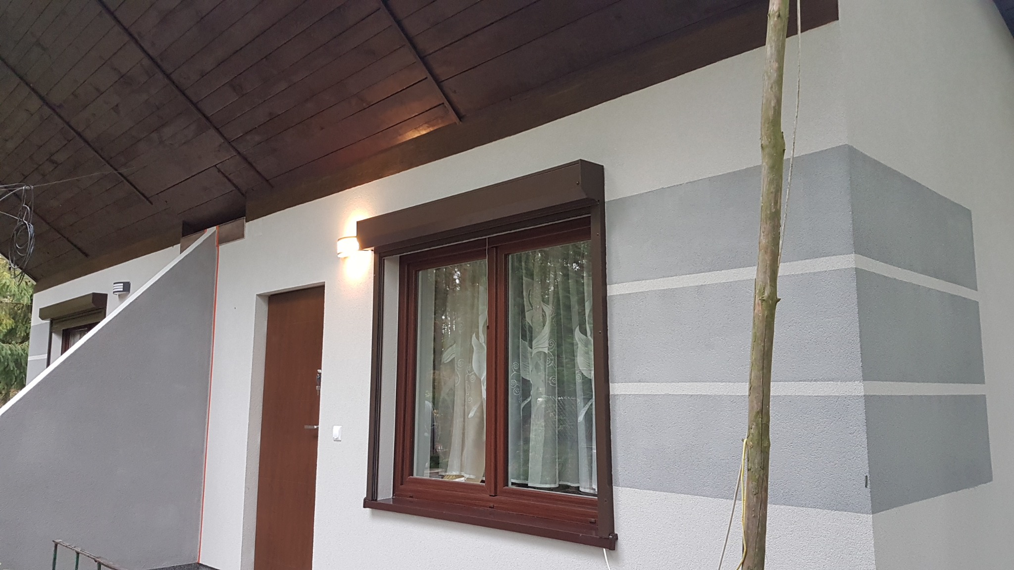 Elewacja domu z oknem z brązowymi roletami i dekoracyjnym malowaniem w poziome pasy szarości. Widoczny fragment dachu i drzwi wejściowe.