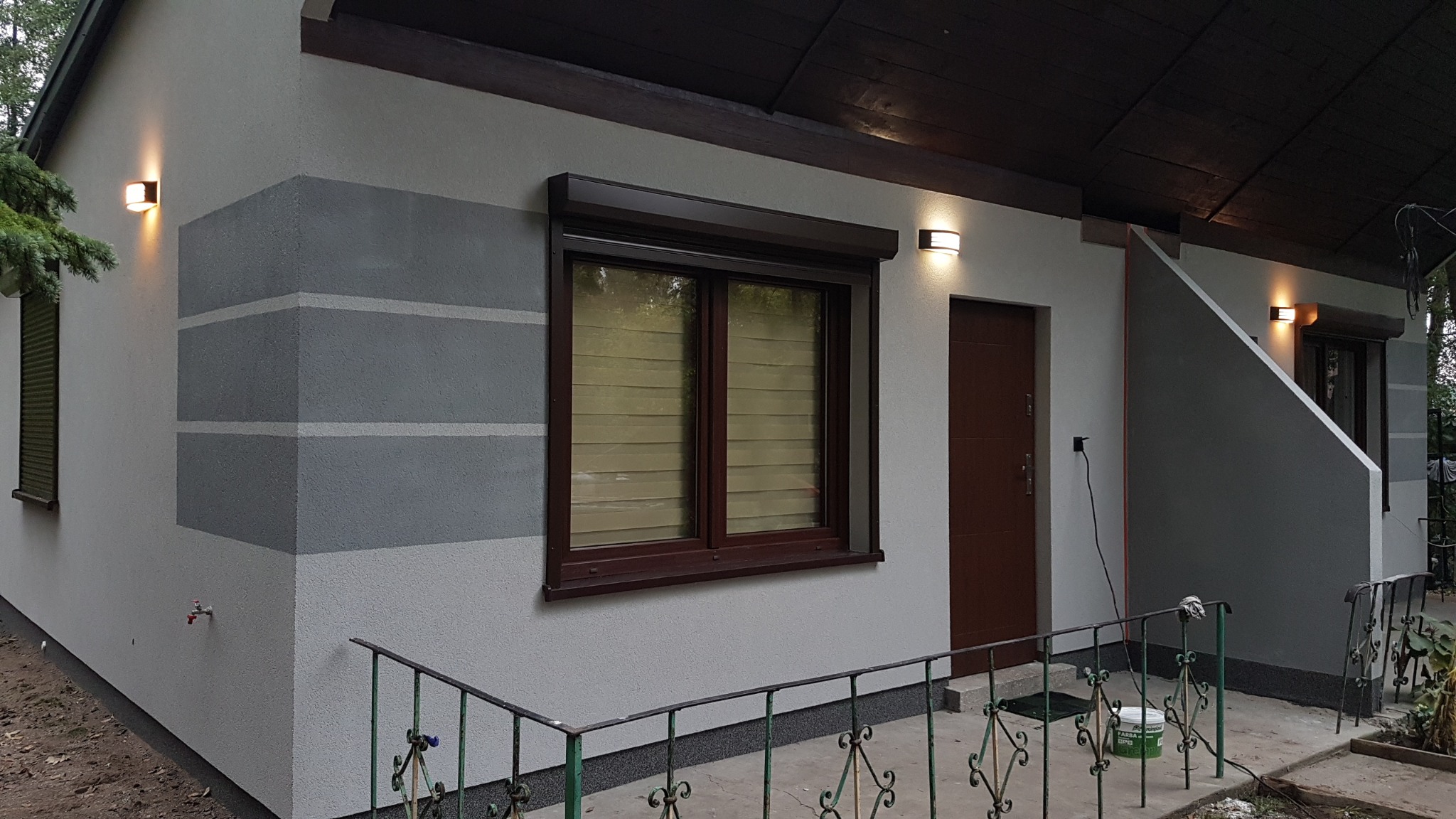Dom po malowaniu elewacji z szarymi pasami, brązowe okna z roletami, oświetlenie zewnętrzne. Widoczne barierki i fragment ogrodu.