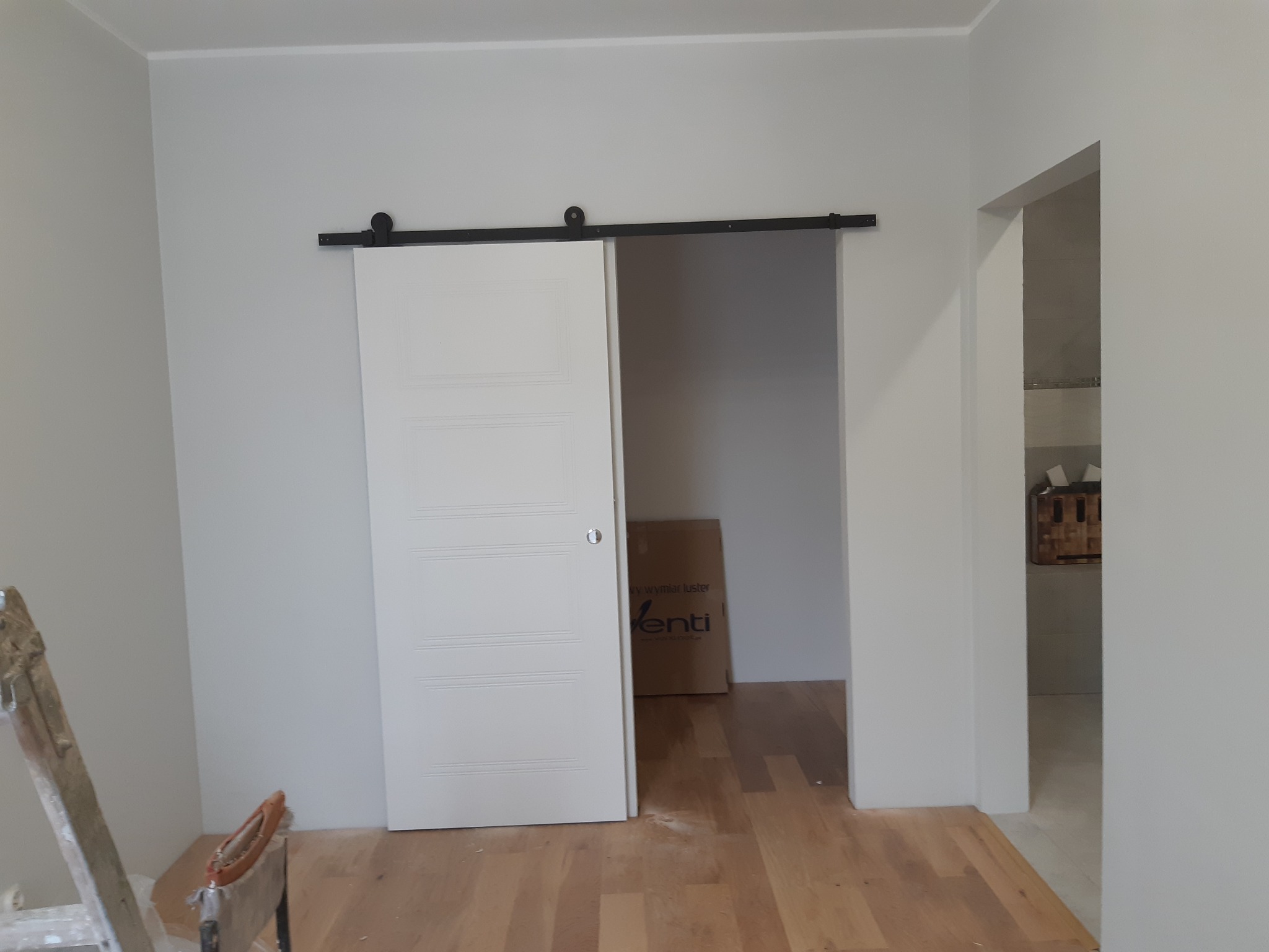Białe, przesuwne drzwi z poziomymi panelami na czarnej szynie, częściowo otwarte, ukazujące wnętrze z drewnianą podłogą i kartonem. Świeżo pomalowane ściany w odcieniach szarości.