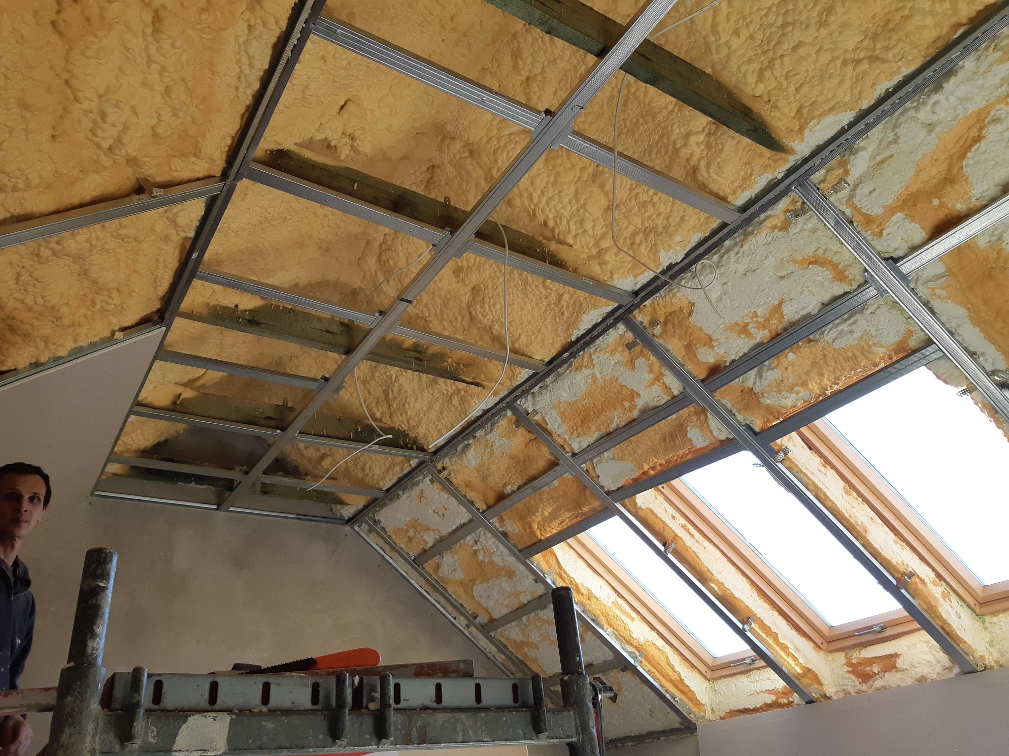 Poddasze z oknem dachowym w trakcie ocieplania pianą PUR, widoczne metalowe profile i fragment rusztowania. Prace izolacyjne w toku.