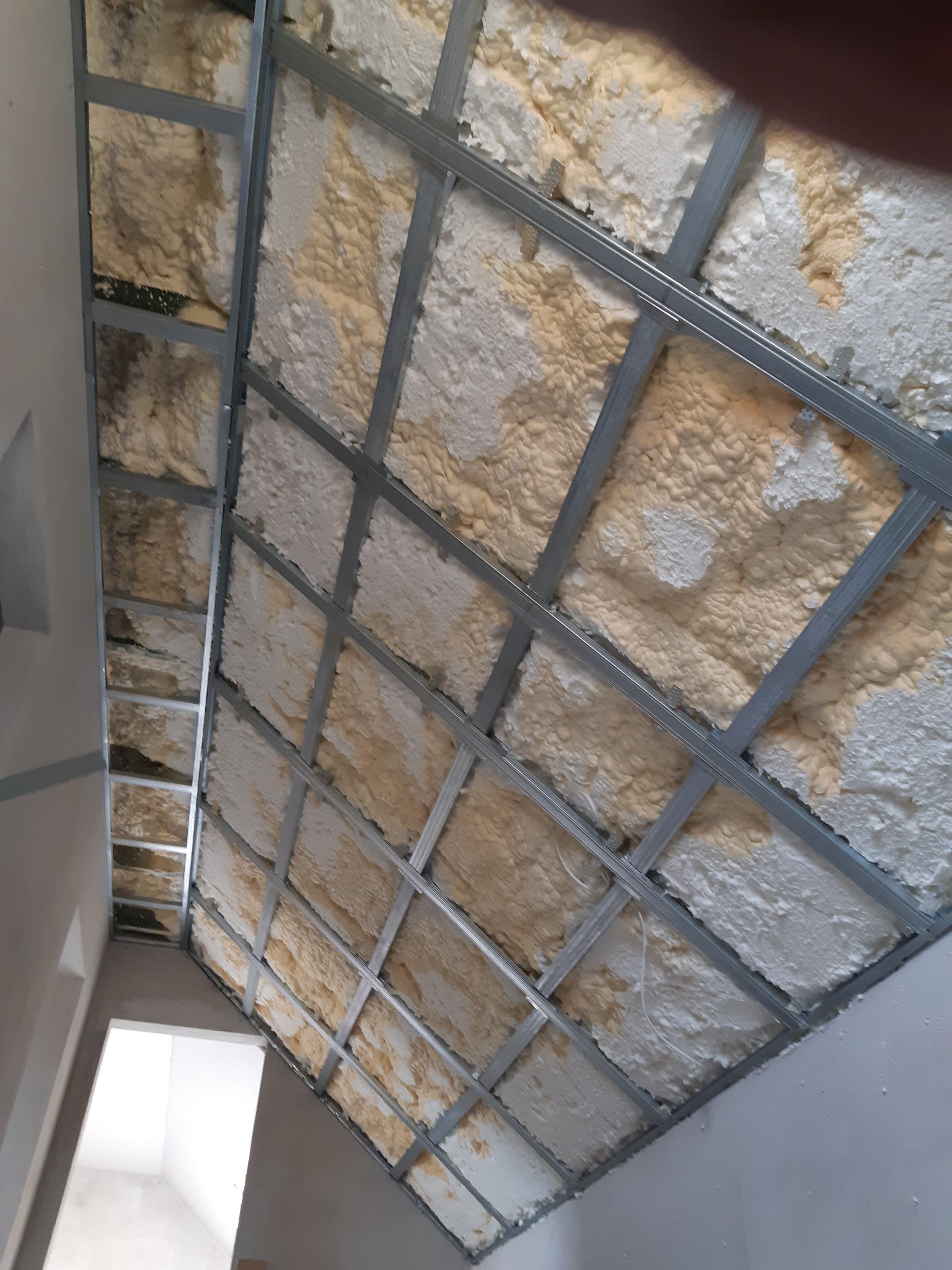 Skośny sufit z metalowym stelażem wypełnionym pianą izolacyjną, widok z dołu, fragment ściany i otworu drzwiowego w tle. Detal ocieplenia poddasza.