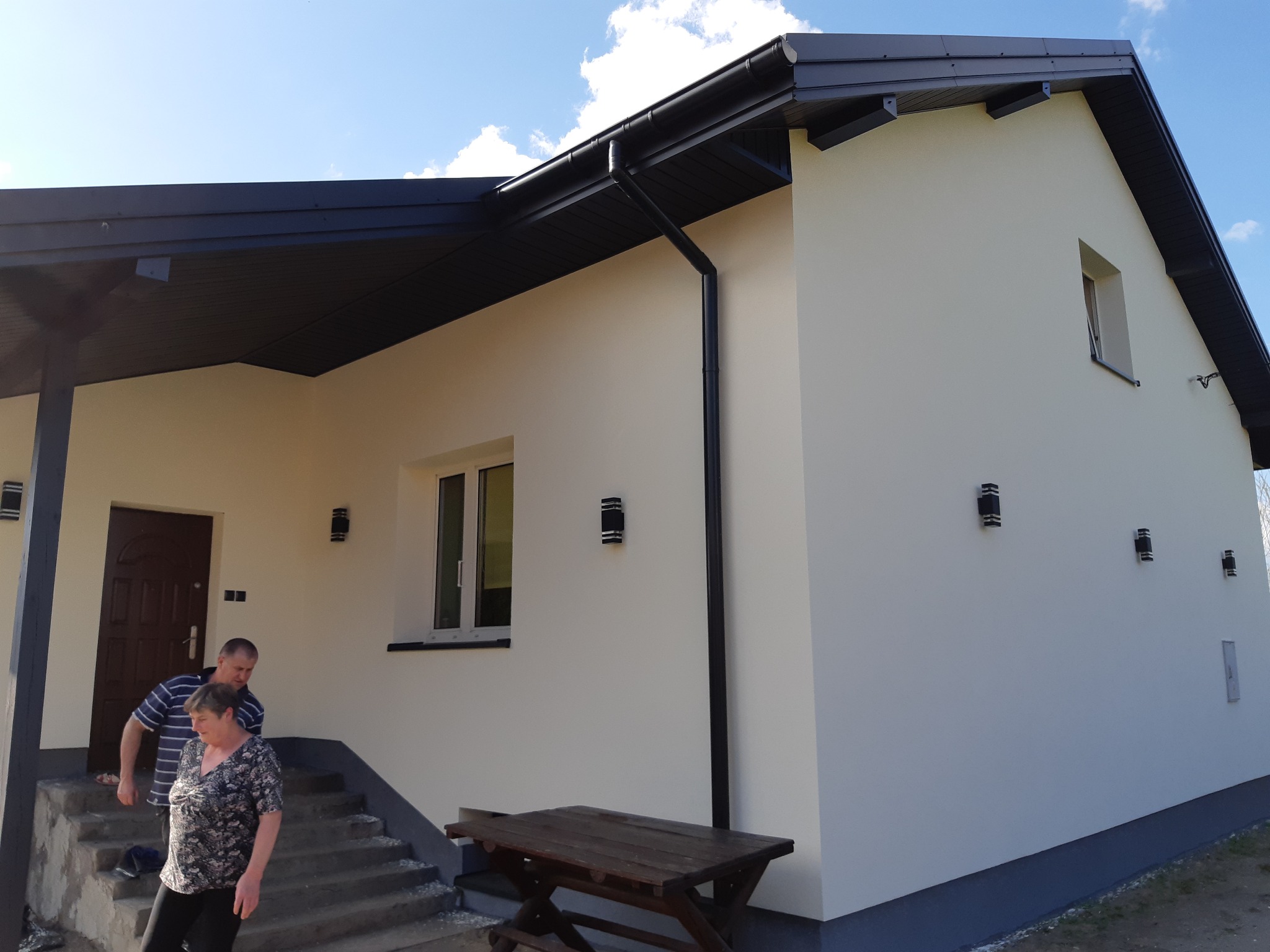 Jasny dom jednorodzinny z czarnym dachem i oświetleniem zewnętrznym, widoczni ludzie na schodach wejściowych. Elewacja w odcieniach bieli i szarości.
