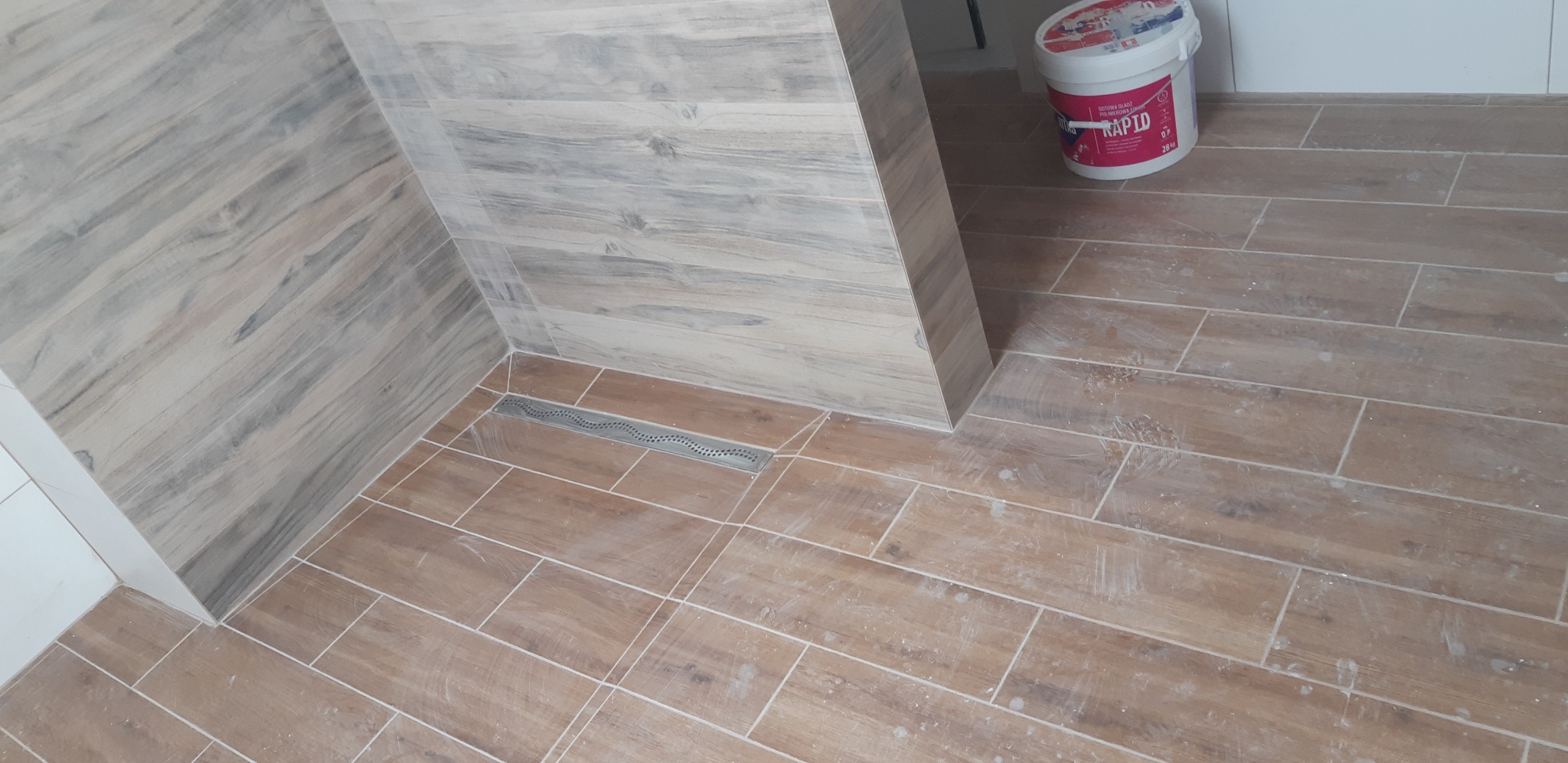 Ukończona łazienka z drewnopodobnymi płytkami, widoczny odpływ liniowy i wiadro z gotową gładzią. Detal wykończenia wnętrza z bliska.