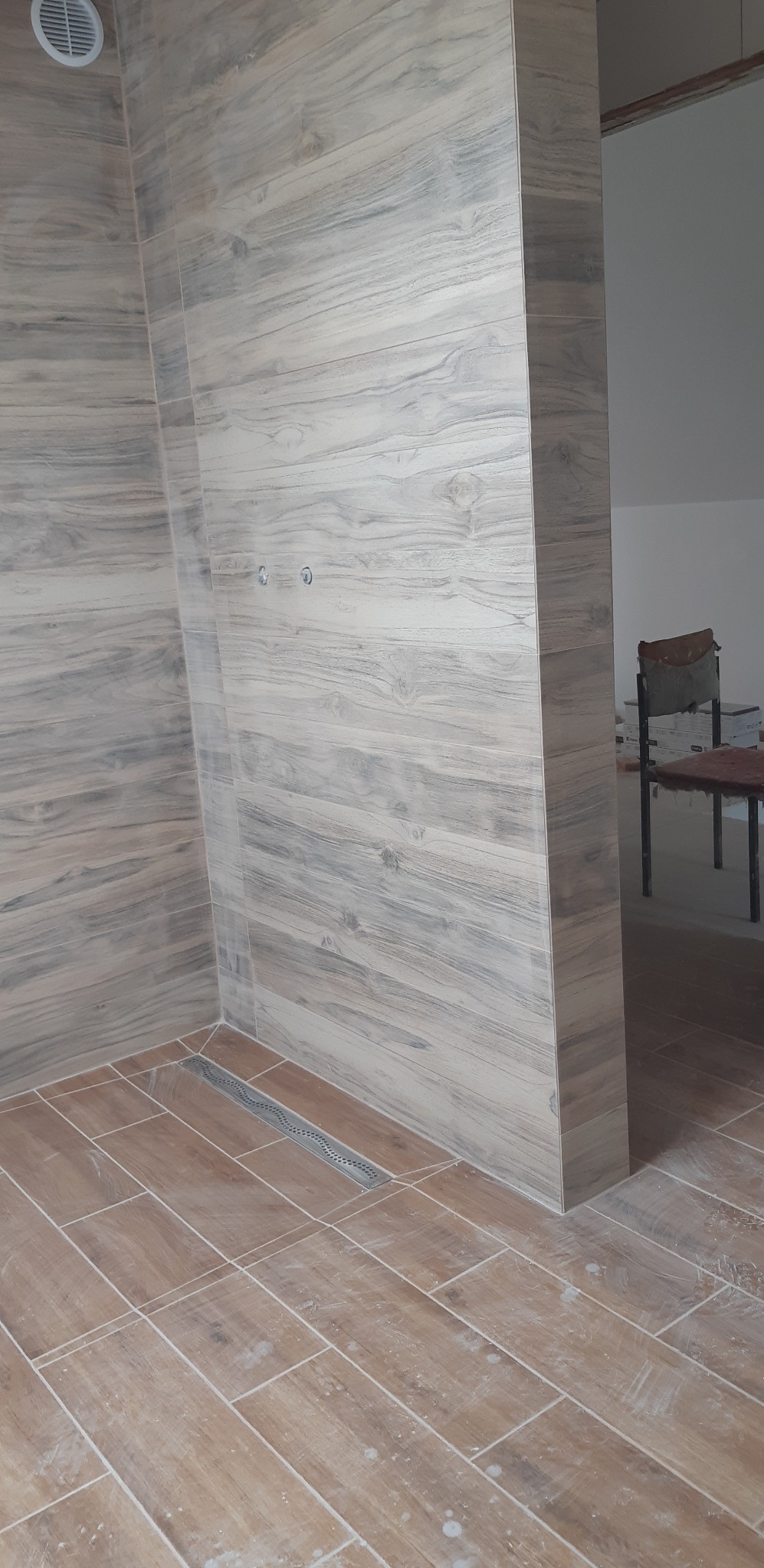 Wykończona łazienka z drewnopodobnymi płytkami na ścianach i podłodze. Widoczny odpływ liniowy i fragment pomieszczenia obok z krzesłem.