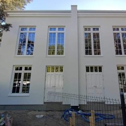 S.A.M.BUD - Biała elewacja dwukondygnacyjnego budynku z symetrycznym układem okien i drzwi, widoczne elementy wykończeniowe i teren wokół w trakcie prac.