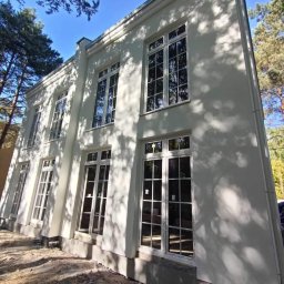 S.A.M.BUD - Biała elewacja dwukondygnacyjnego budynku z dużymi oknami z białymi szprosami, w otoczeniu drzew i z ziemią w stanie surowym u podstawy.