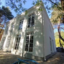 S.A.M.BUD - Dwupiętrowy dom z białymi ścianami i dużymi oknami, otoczony drzewami. Widoczne rury na ziemi sugerują prace wykończeniowe.