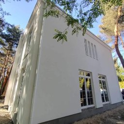 S.A.M.BUD - Biały, dwukondygnacyjny dom z oknami w stylu retro, otoczony drzewami. Świeżo otynkowana elewacja w kontraście z naturalnym otoczeniem. Ujęcie z dołu.