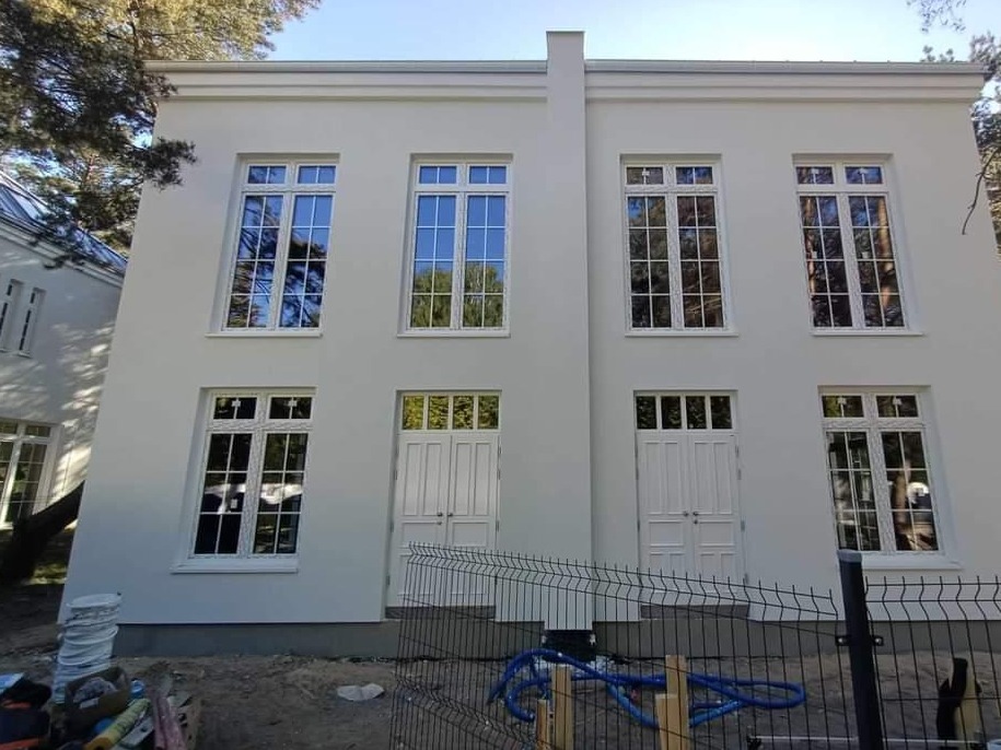 Biała elewacja dwukondygnacyjnego budynku z symetrycznym układem okien i drzwi, widoczne elementy wykończeniowe i teren wokół w trakcie prac.