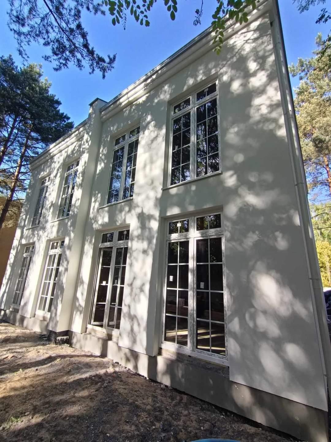 Biała elewacja dwukondygnacyjnego budynku z dużymi oknami z białymi szprosami, w otoczeniu drzew i z ziemią w stanie surowym u podstawy.