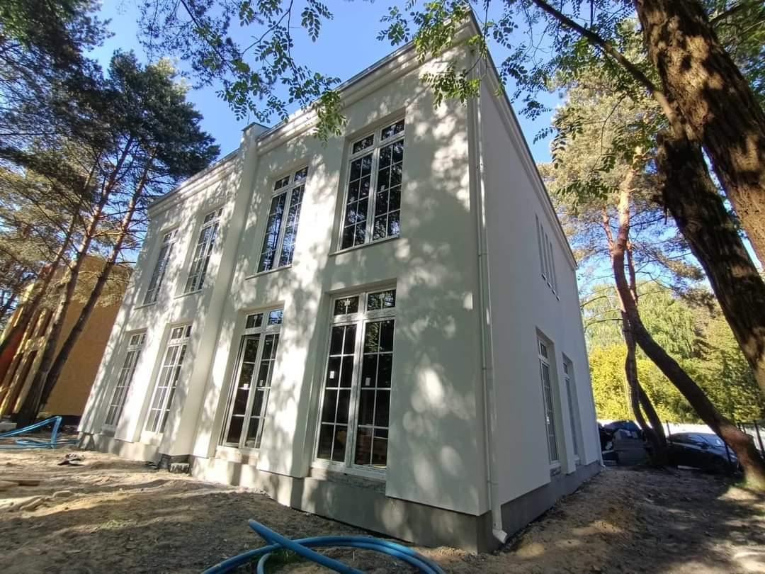 Dwupiętrowy dom z białymi ścianami i dużymi oknami, otoczony drzewami. Widoczne rury na ziemi sugerują prace wykończeniowe.