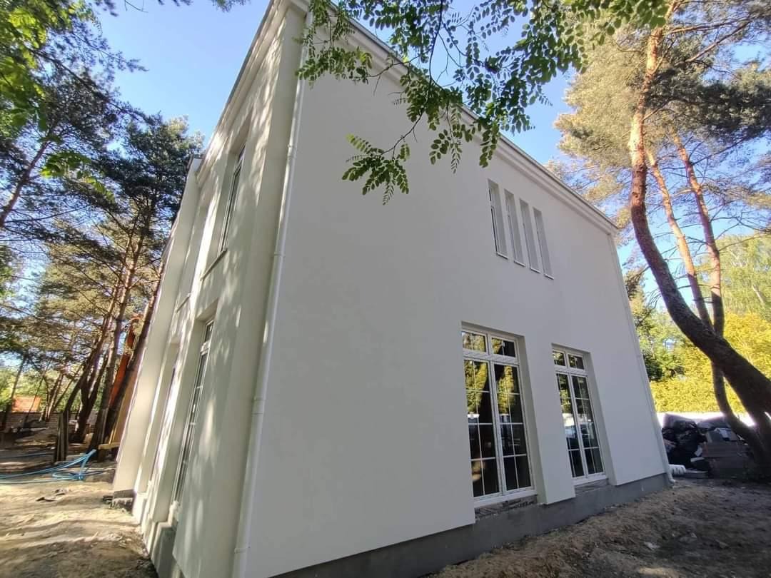 Biały, dwukondygnacyjny dom z oknami w stylu retro, otoczony drzewami. Świeżo otynkowana elewacja w kontraście z naturalnym otoczeniem. Ujęcie z dołu.