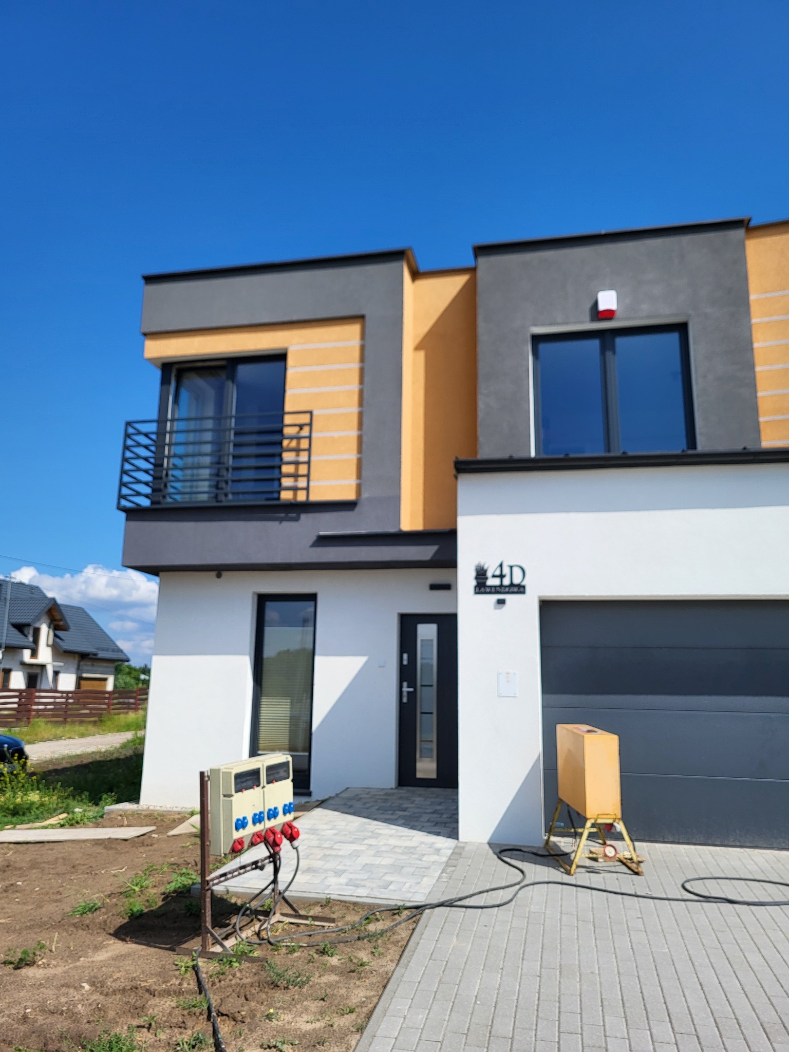 Dwukondygnacyjny dom z płaskim dachem, wykończony w bieli, szarości i odcieniach pomarańczy. Widoczny balkon z czarną balustradą i skrzynka elektryczna na pierwszym planie.