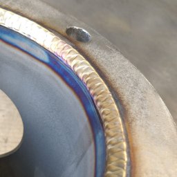 Welding and Metalworks Sebastian Piekarczyk - Szczegółowe zbliżenie na spawany element metalowy z widocznym, precyzyjnym spoiwem i opalizującymi przebarwieniami termicznymi. Widoczny otwór montażowy.
