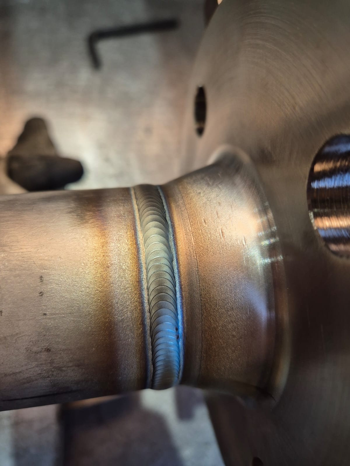 Precyzyjne spoiny na metalowym elemencie, widoczne detale i kolory powstałe w procesie łączenia metali. Ślady obróbki na powierzchni.