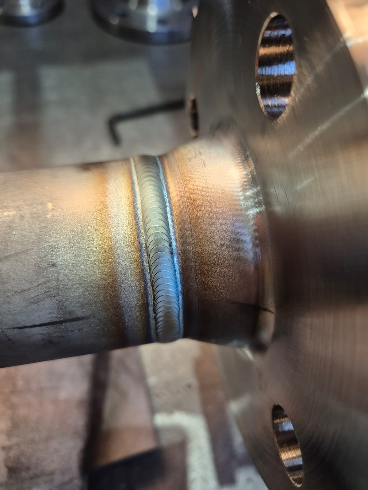 Precyzyjne połączenie spawane rury z kołnierzem, widoczne regularne spoiny i przebarwienia termiczne na metalu. Detal z bliska, oświetlenie warsztatowe.