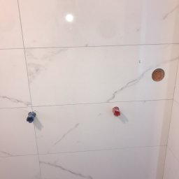 Dmytrów Andrzej - Ściana wyłożona białymi płytkami imitującymi marmur z trzema wystającymi rurami (czerwona, niebieska i miedziana) przygotowanymi do podłączenia armatury.