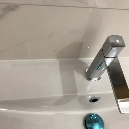 Dmytrów Andrzej - Bateria umywalkowa, chromowana, o nowoczesnym, geometrycznym kształcie, zamontowana na białej umywalce z marmurowym tłem. Widoczny korek umywalki.