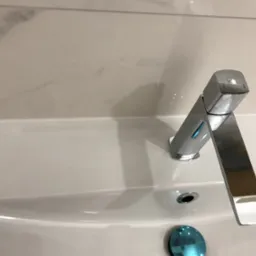 Bateria umywalkowa, chromowana, o nowoczesnym, geometrycznym kształcie, zamontowana na białej umywalce z marmurowym tłem. Widoczny korek umywalki.