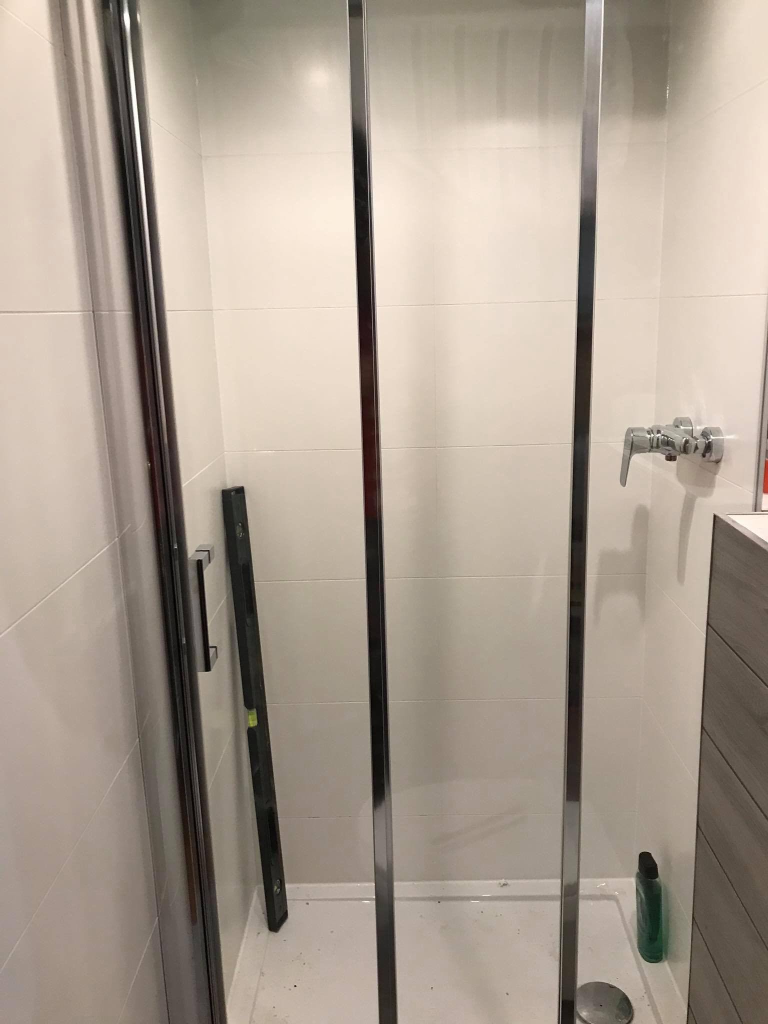 Kabina prysznicowa z chromowanymi profilami i poziomica wewnątrz, białe płytki na ścianach, bateria umywalkowa. Widok na wnętrze kabiny.