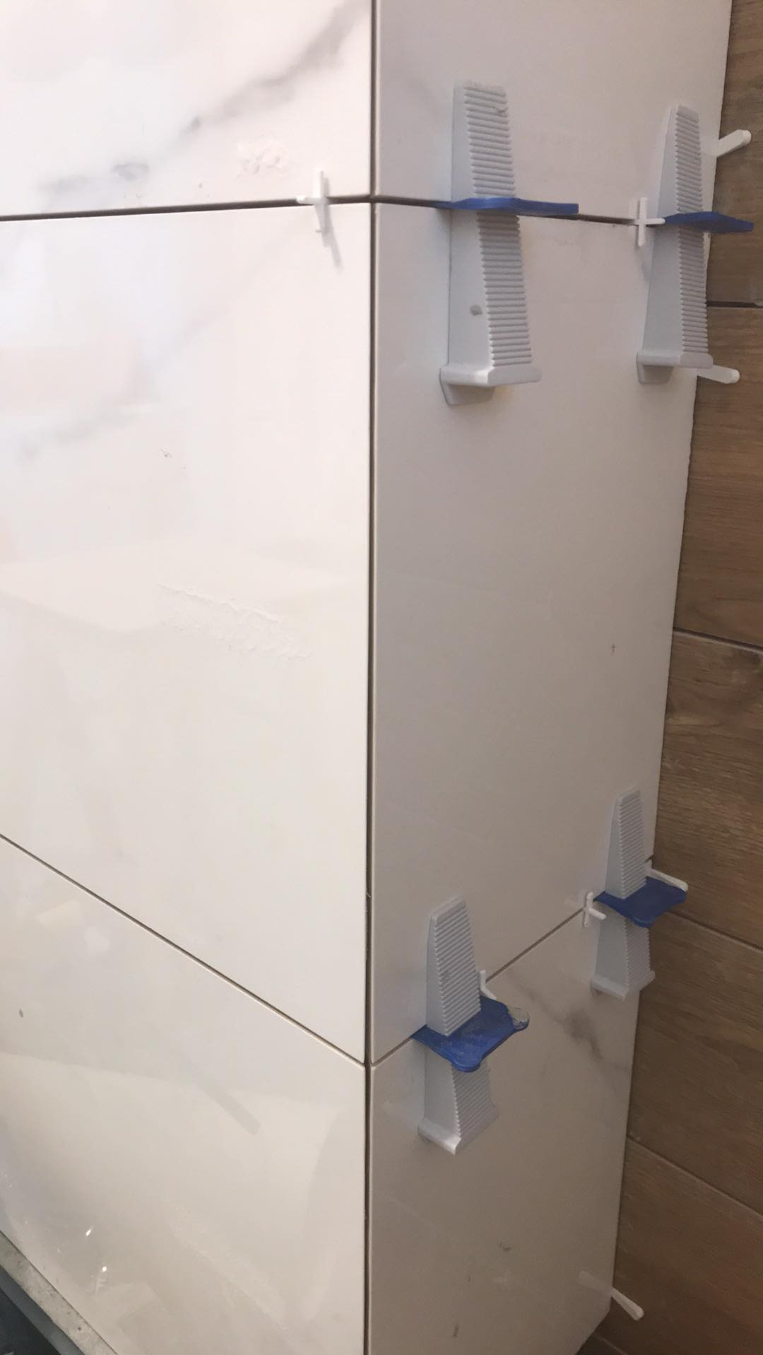 Białe płytki imitujące marmur, ułożone z użyciem systemu poziomowania płytek, widoczne krzyżyki dystansowe i kliny. Fragment ściany z płytek drewnopodobnych w tle.