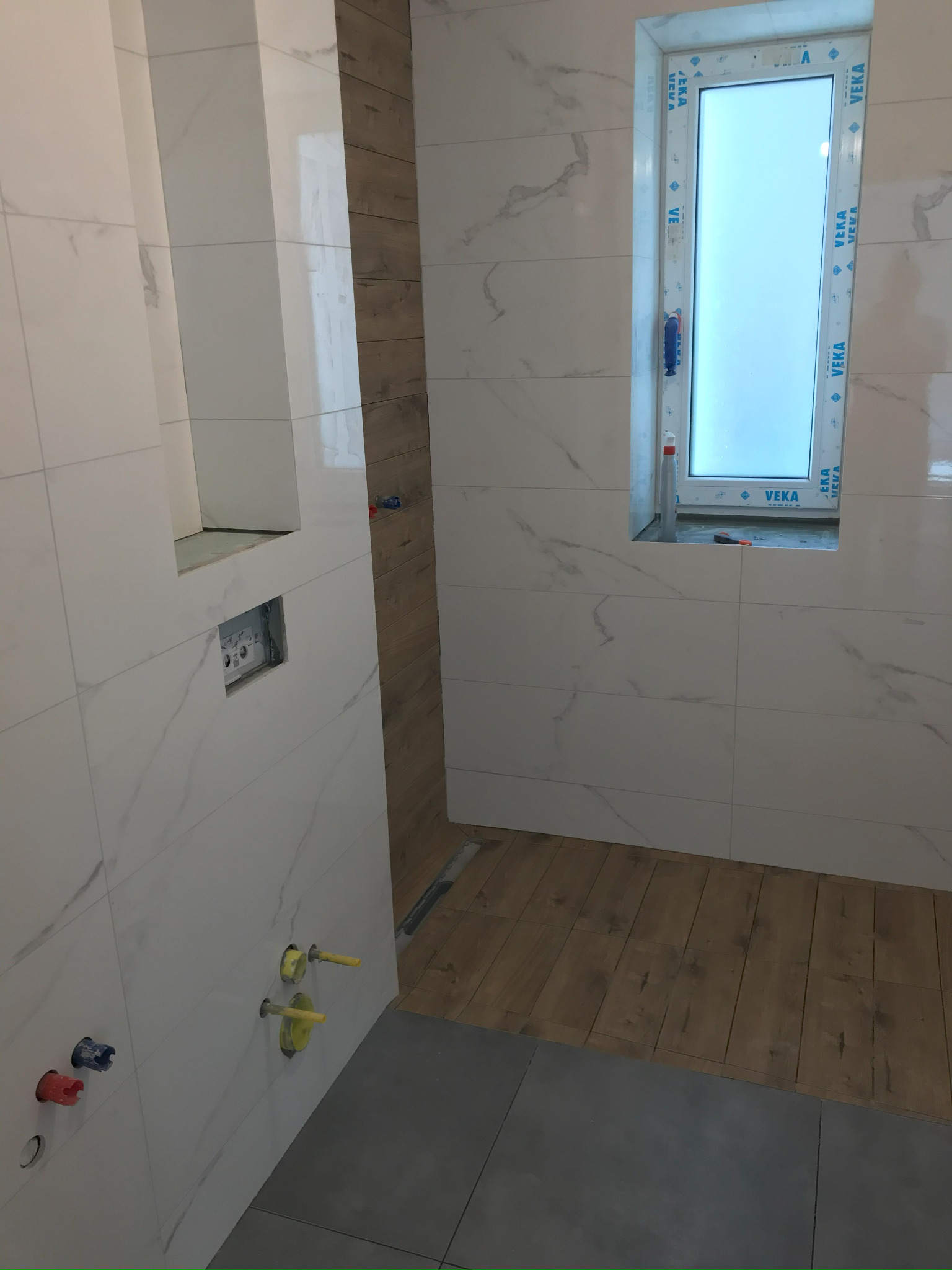 Wykończona łazienka z oknem: białe płytki imitujące marmur na ścianach, drewnopodobne panele i szara podłoga. Widoczne instalacje wodne i okno PCV.