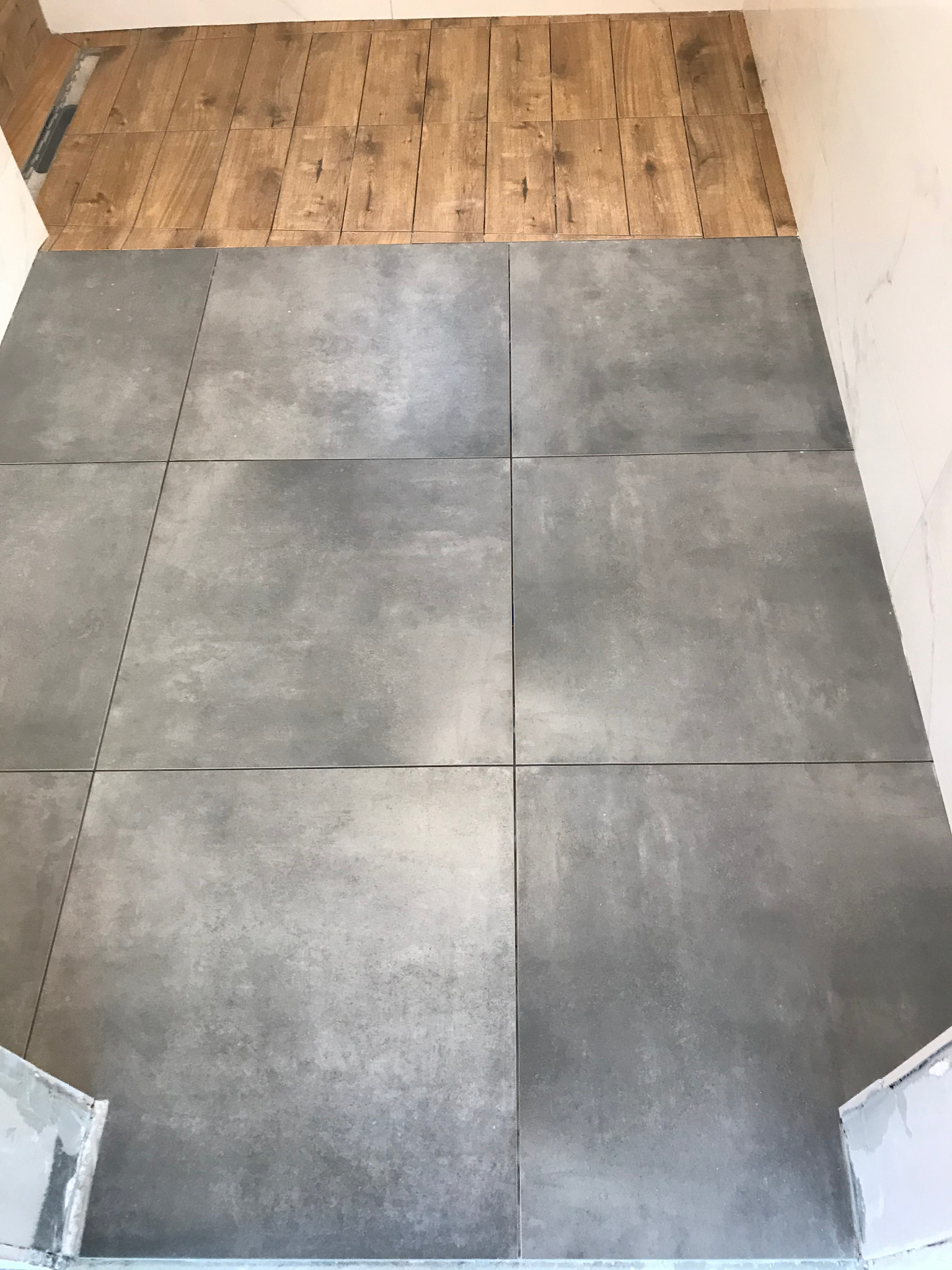 Ułożone szare płytki podłogowe w łazience, z widocznym fragmentem drewnopodobnej podłogi i białych ścian. Widok z góry na świeżo wykonaną posadzkę.