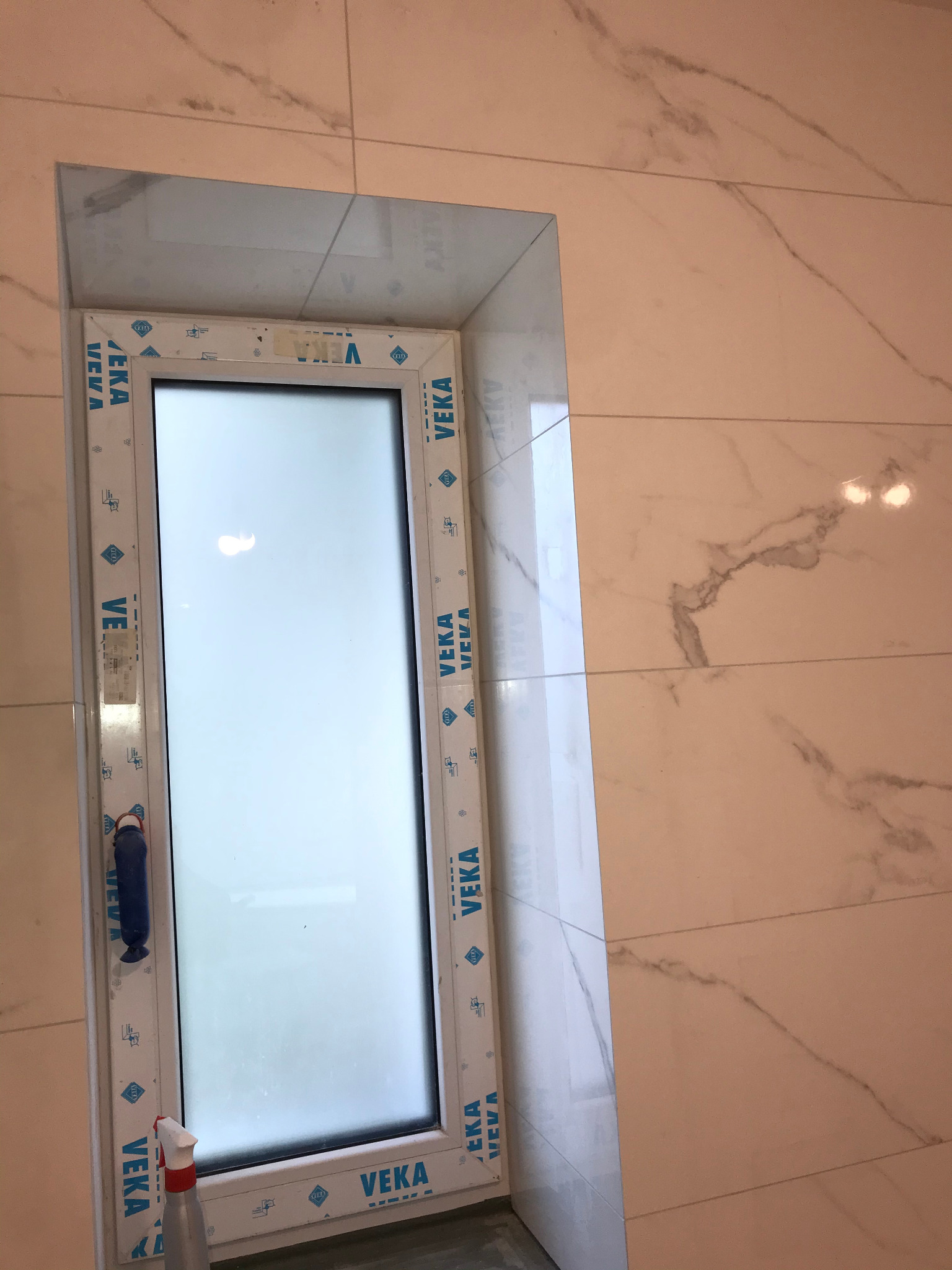 Wnęka okienna w łazience wykończona płytkami imitującymi marmur. Białe okno PCV marki Veka. Widoczne narzędzia glazurnicze.