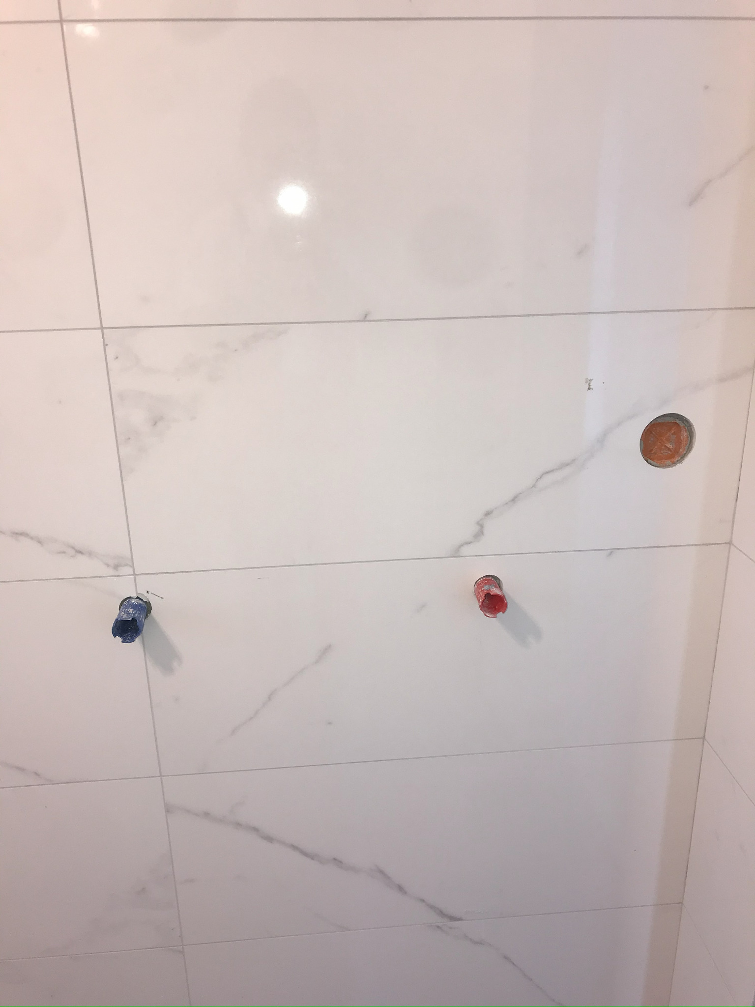 Ściana wyłożona białymi płytkami imitującymi marmur z trzema wystającymi rurami (czerwona, niebieska i miedziana) przygotowanymi do podłączenia armatury.