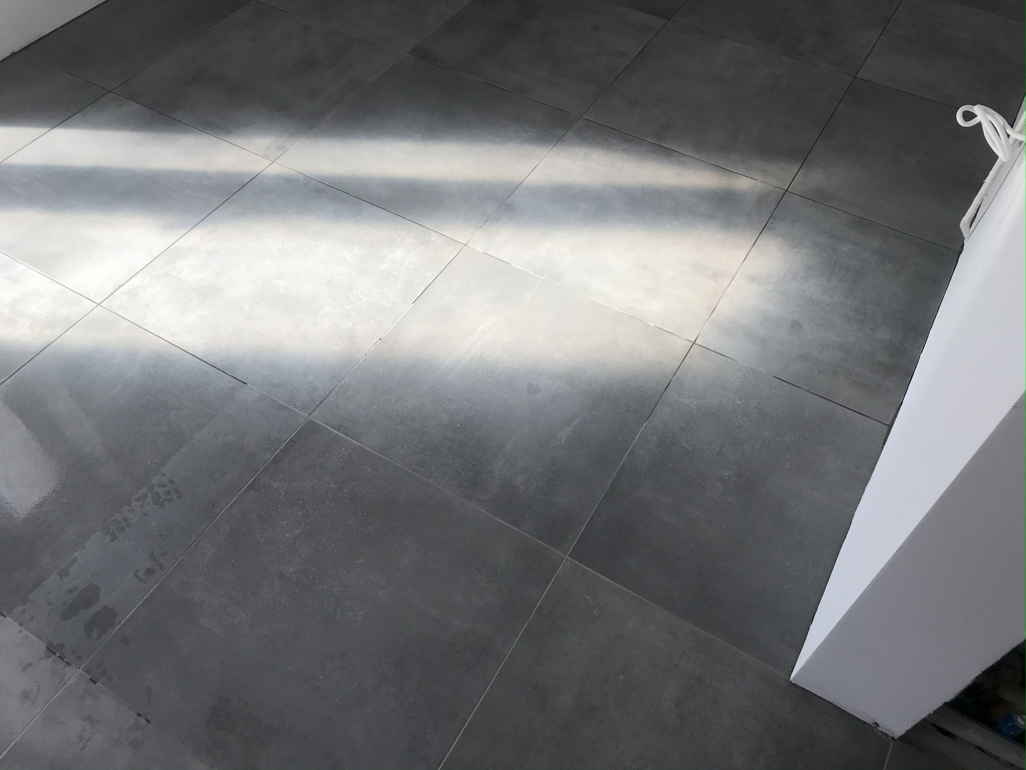 Szare, matowe płytki podłogowe ułożone w kwadraty, oświetlone naturalnym światłem. Fragment białej ściany w prawym dolnym rogu kadru. Minimalistyczny design.