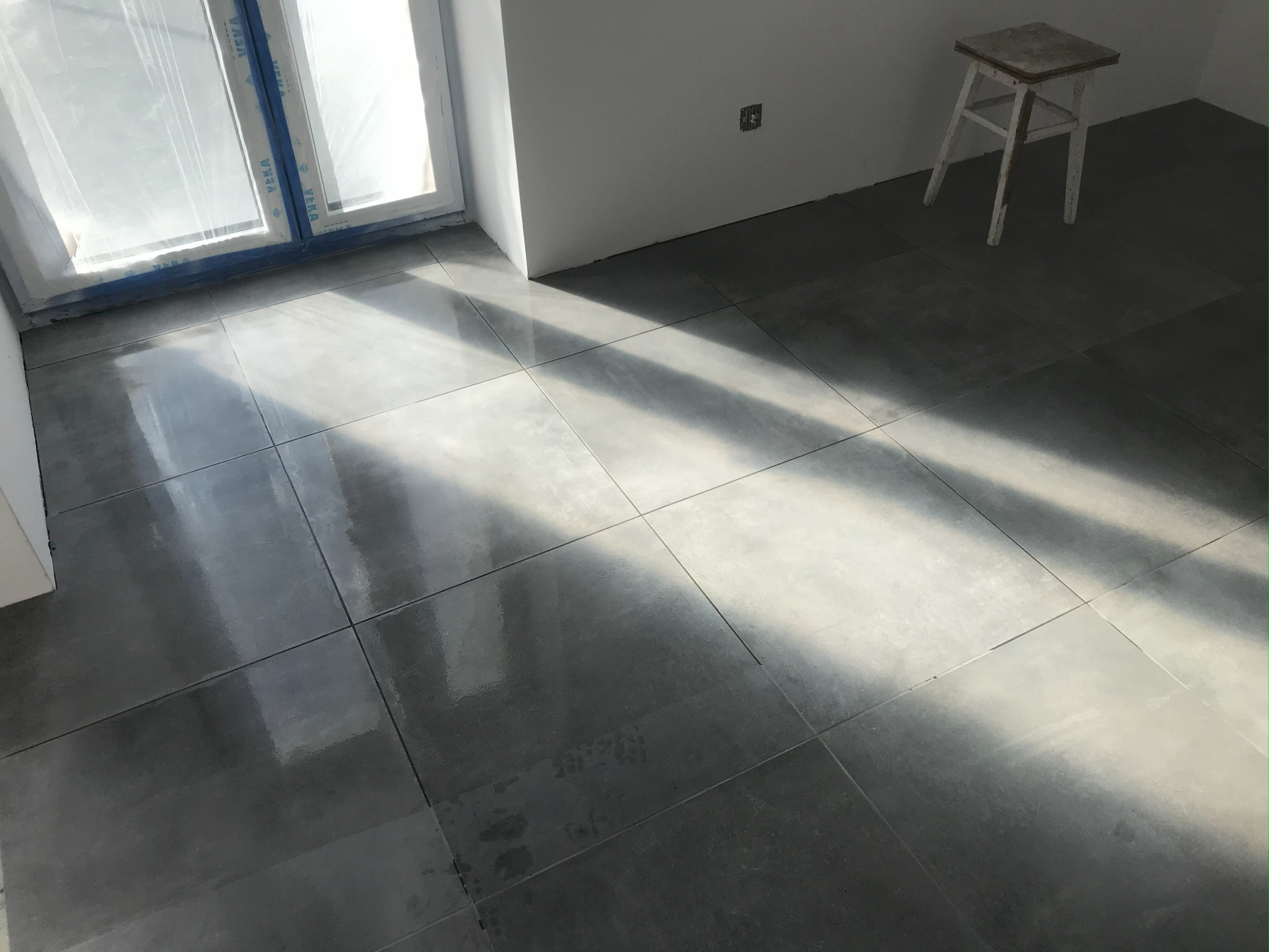 Ułożone szare płytki podłogowe w słonecznym pokoju. Okno zabezpieczone folią malarską. Biały taboret w rogu. Widok na świeżo wykafelkowaną podłogę.