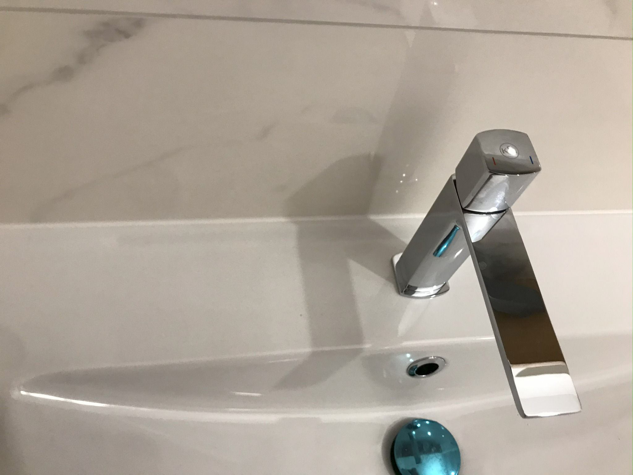 Bateria umywalkowa, chromowana, o nowoczesnym, geometrycznym kształcie, zamontowana na białej umywalce z marmurowym tłem. Widoczny korek umywalki.